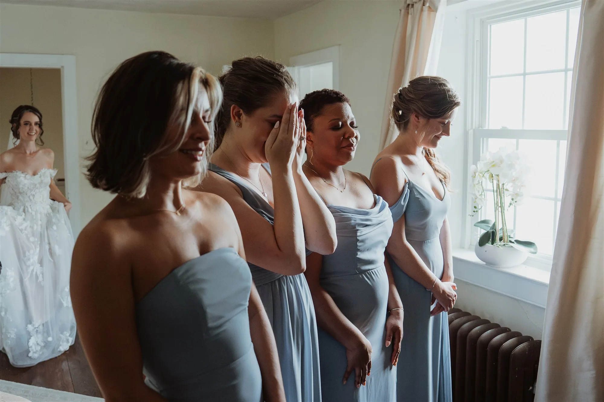 Four bridesmaids in dusty blue gowns react with tears seeing the bride for the first time