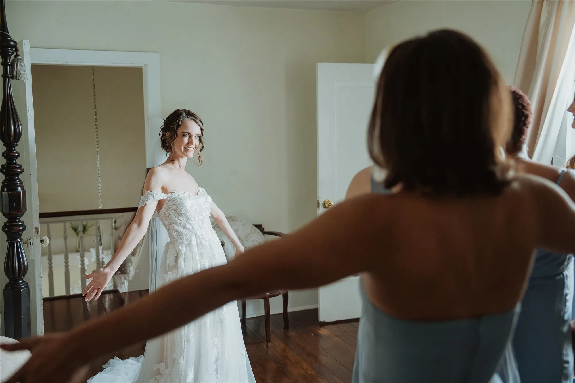 Bride in lace gown smiles as bridesmaid reaches toward her during getting-ready moment inside Rixey Manor