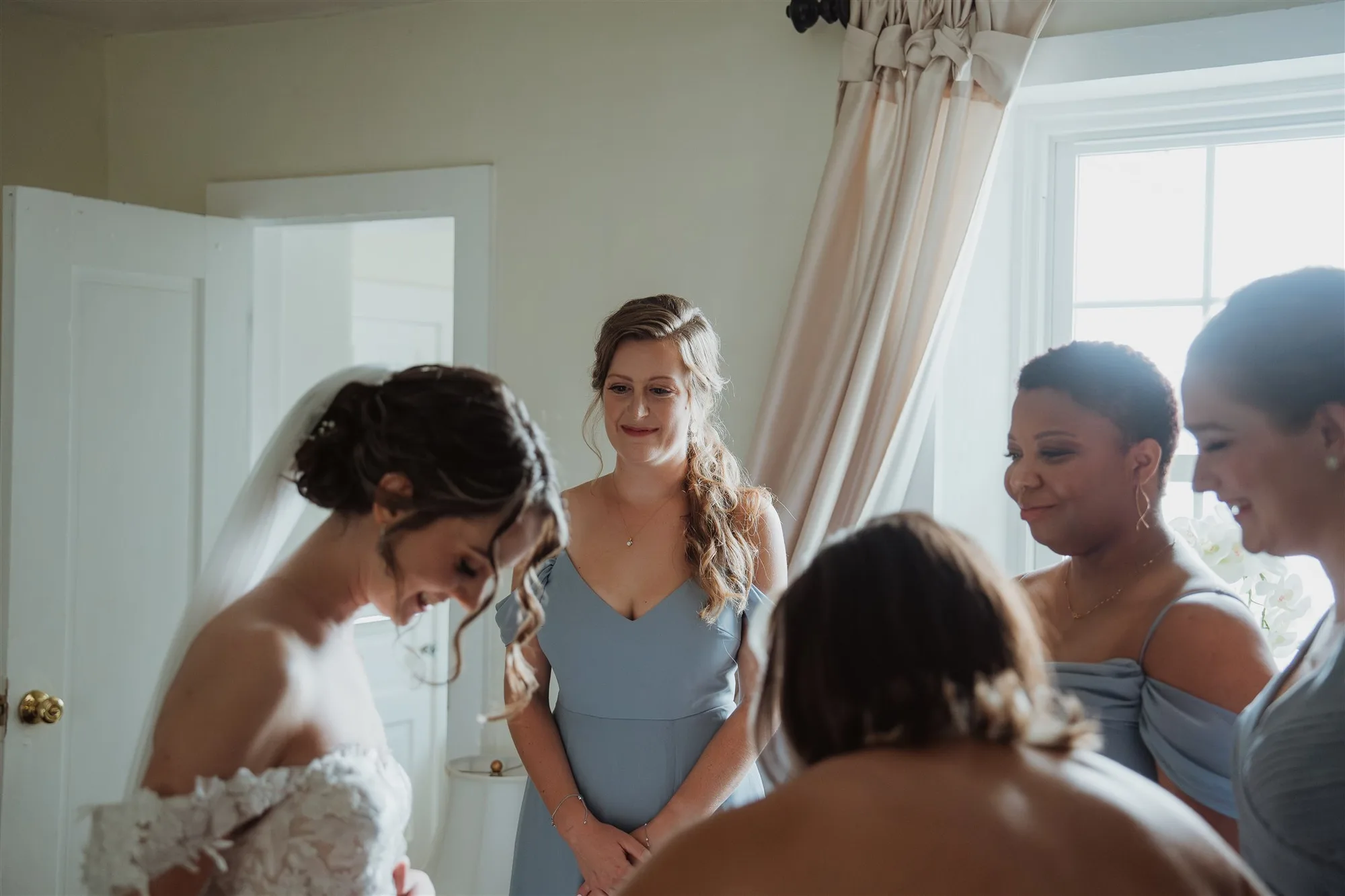 Bride gets ready as smiling bridesmaids in blue dresses gather around her in a bright dressing room