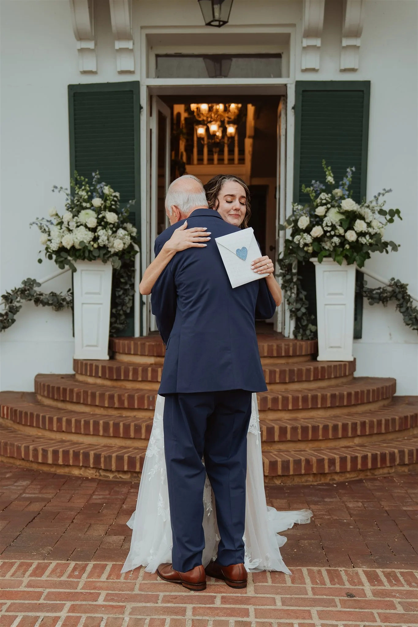 Bride embraces older gentleman on Rixey Manor's brick carriage steps, flanked by white floral arrangements