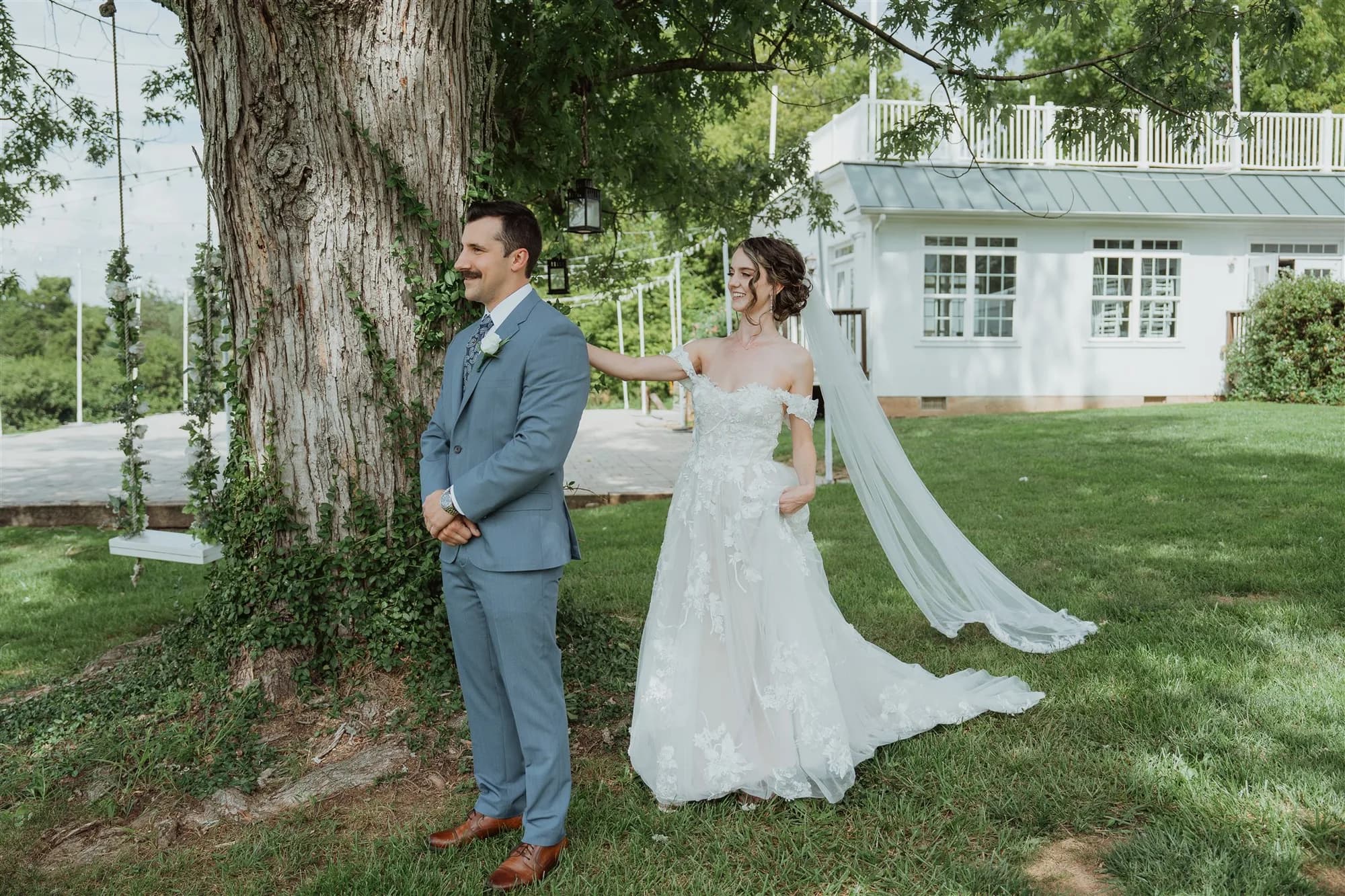 Bride taps groom's shoulder during first look under large tree on Rixey Manor grounds, white estate visible behind