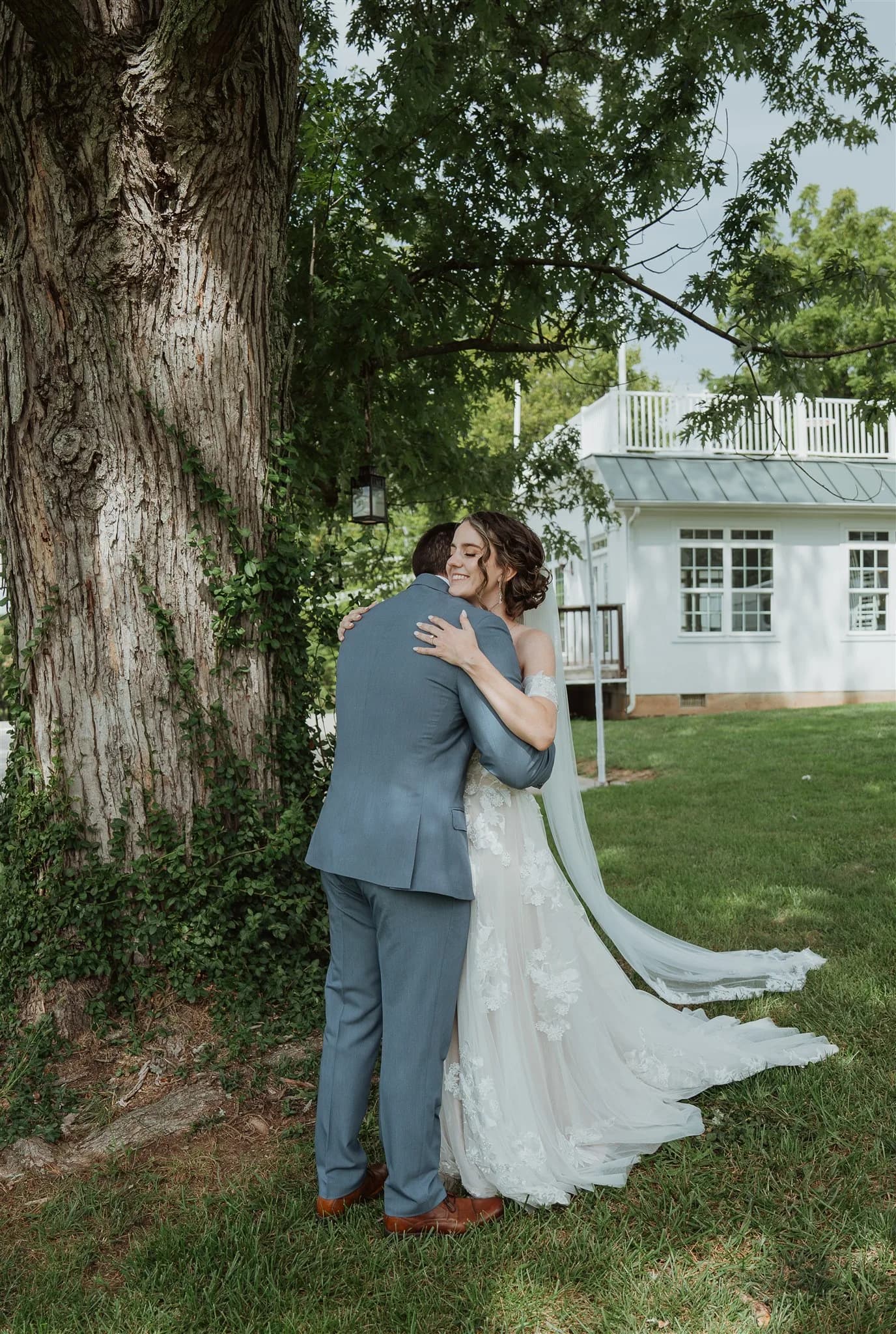 Bride smiling joyfully as she embraces groom during first look beneath large tree on Rixey Manor grounds
