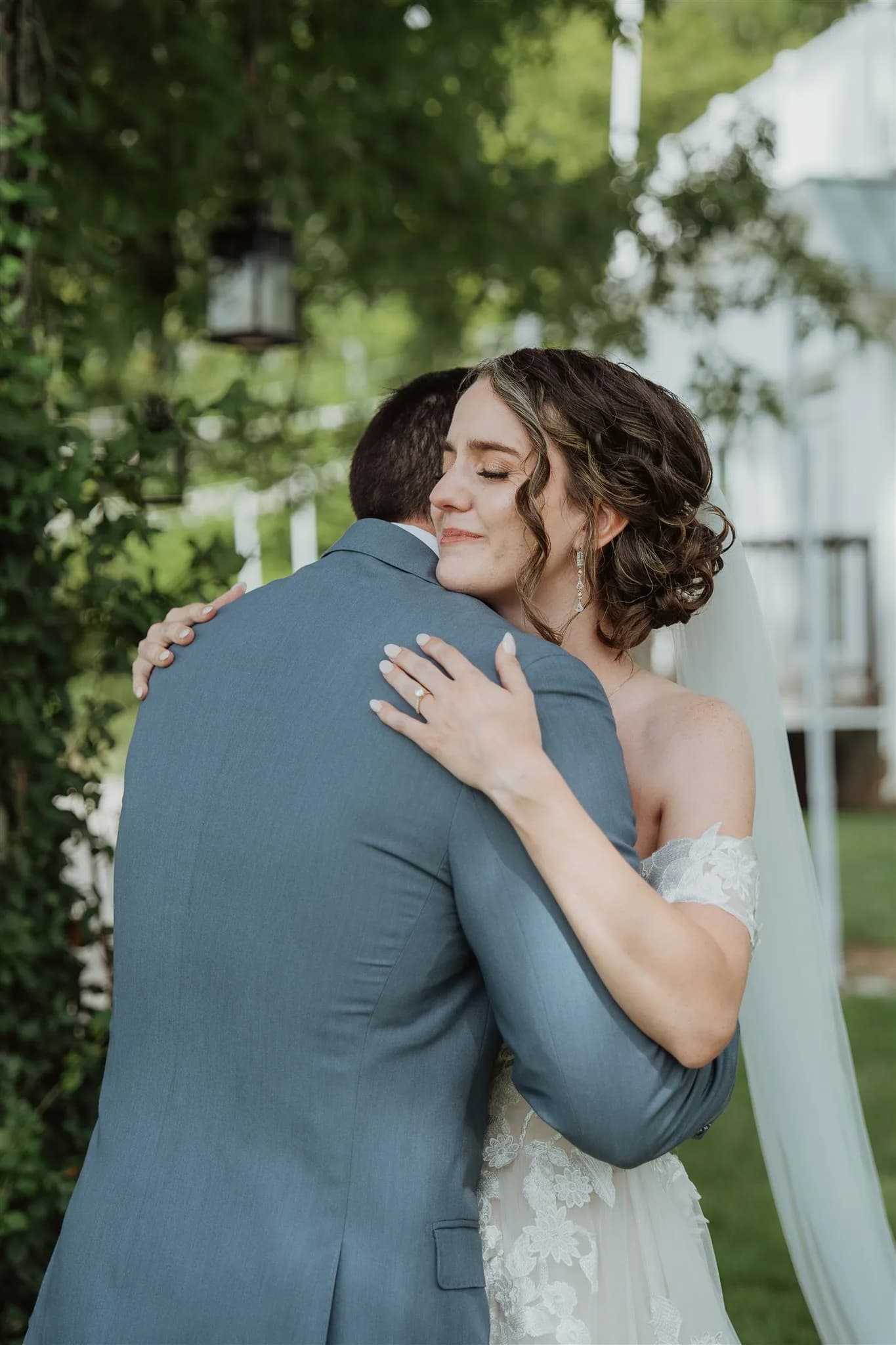 Bride embraces groom during first look, eyes closed in a peaceful smile, surrounded by lush greenery