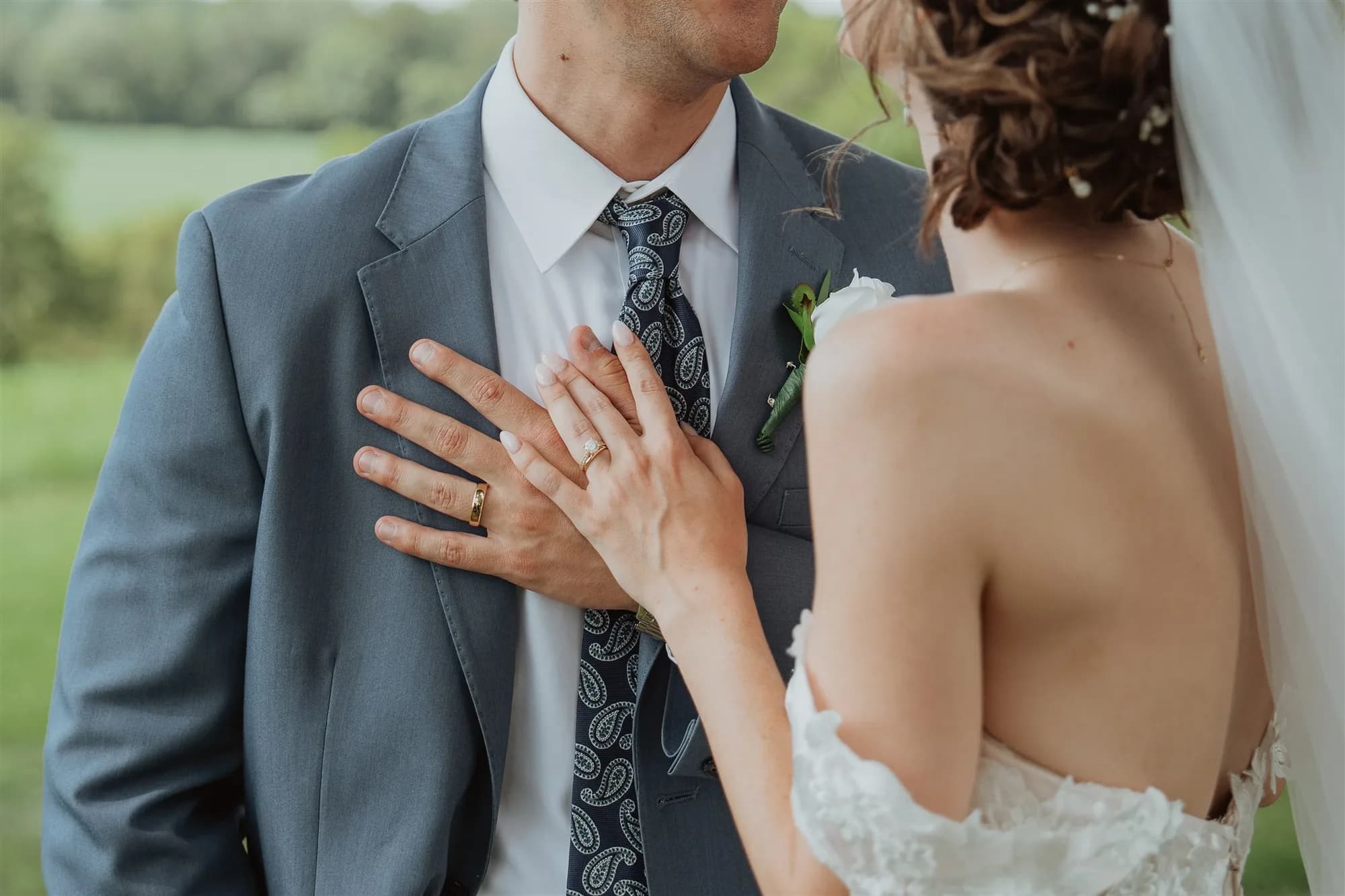 Bride places hand on groom's chest showing wedding rings, both in wedding attire with green landscape behind