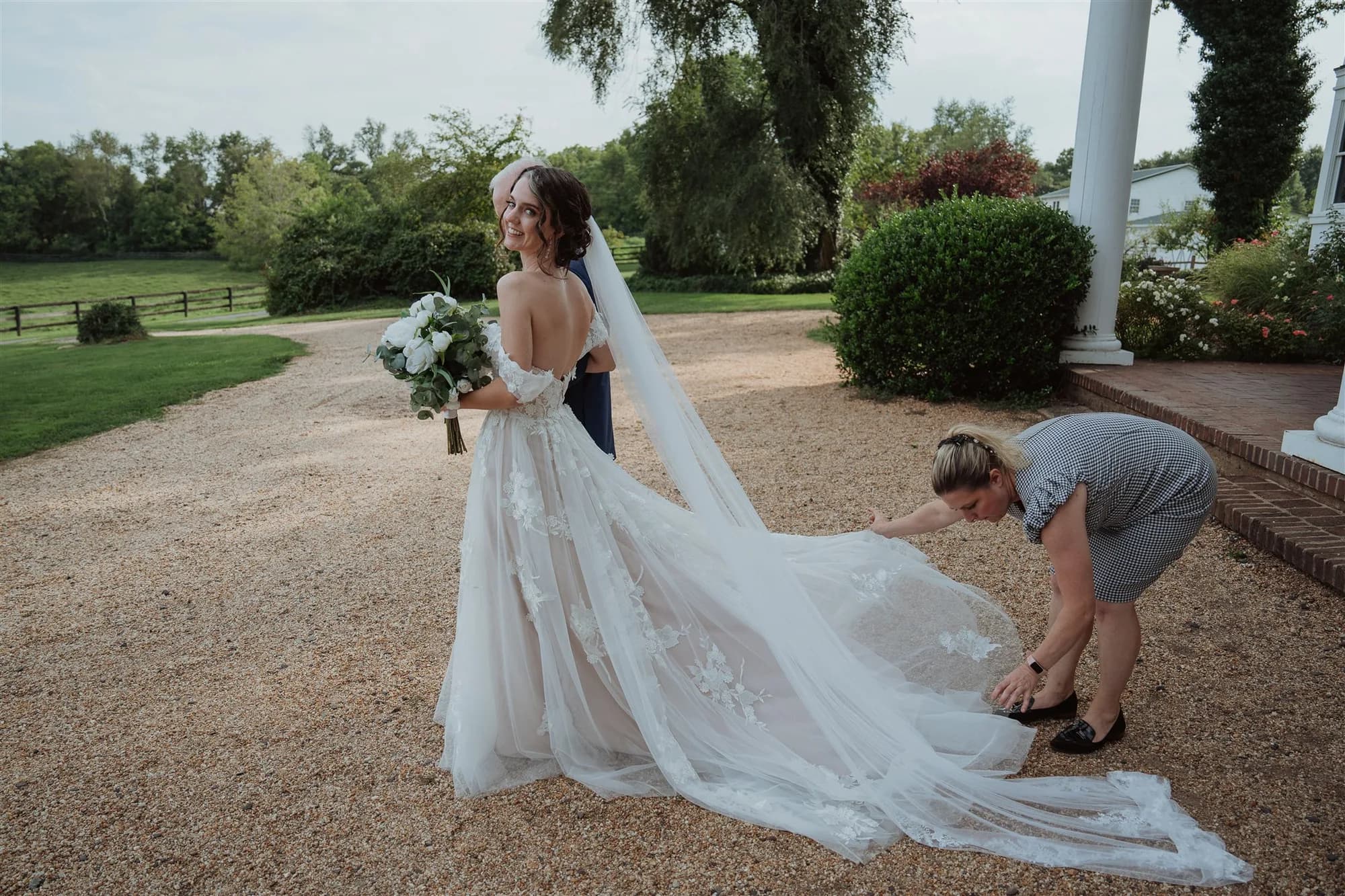 Bride smiles as attendant arranges gown train on Rixey Manor's gravel drive, groom embracing her from behind