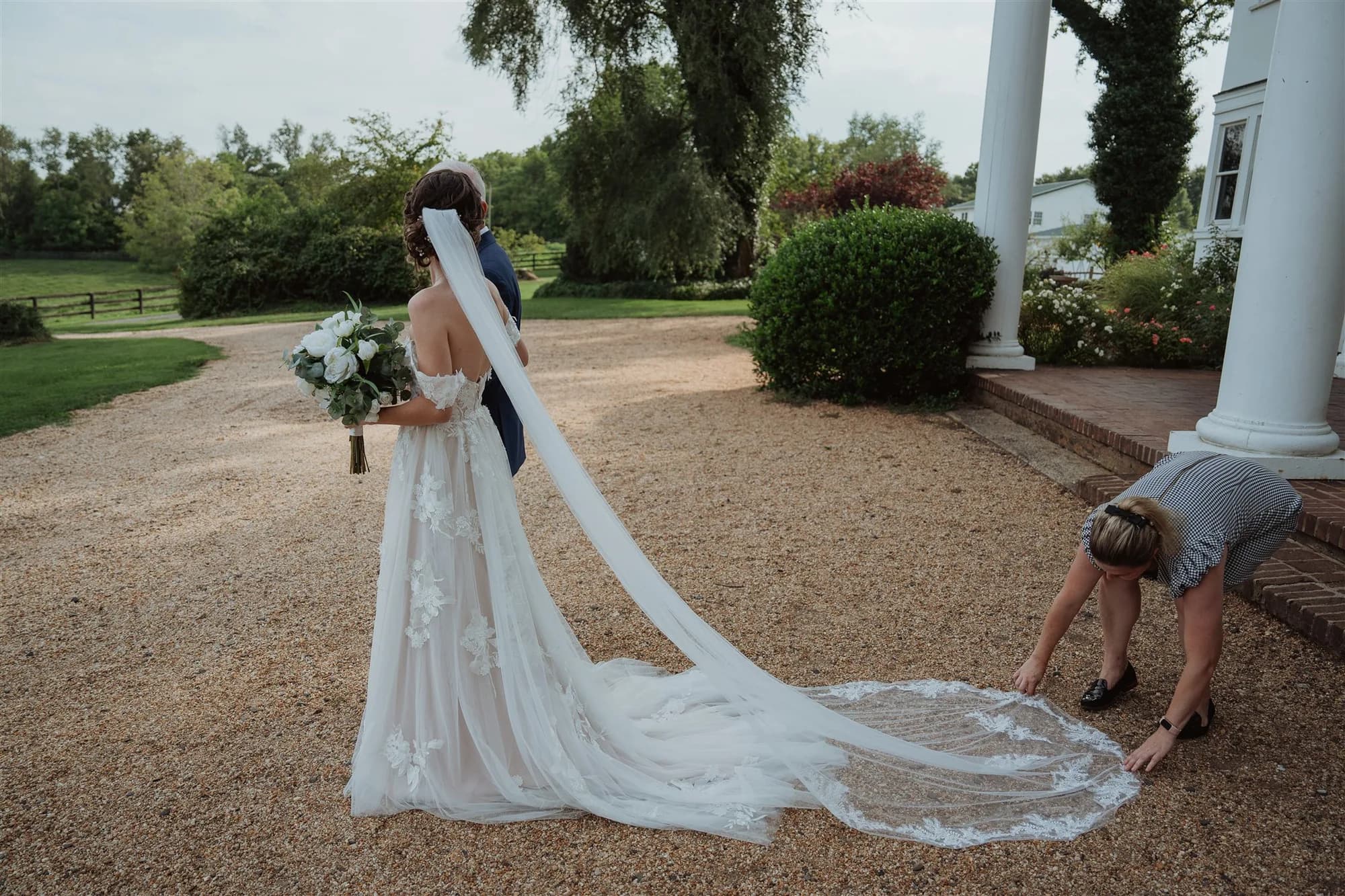 Bride and groom stand on gravel drive at Rixey Manor as assistant arranges cathedral veil train