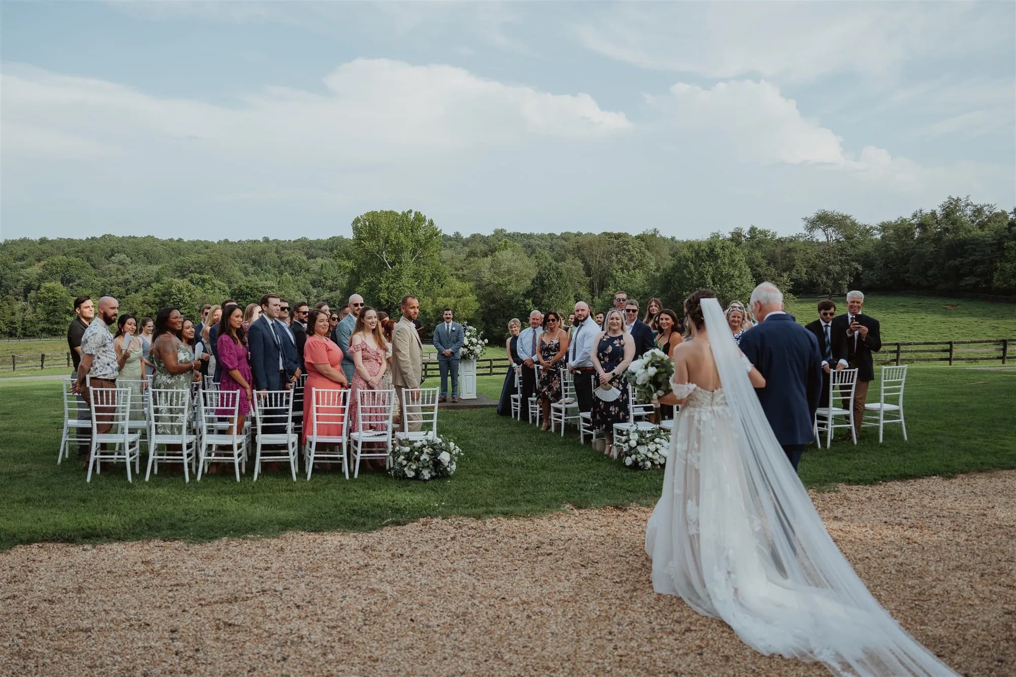 Bride and escort walk the outdoor aisle at Rixey Manor as guests stand for the ceremony on lush green grounds