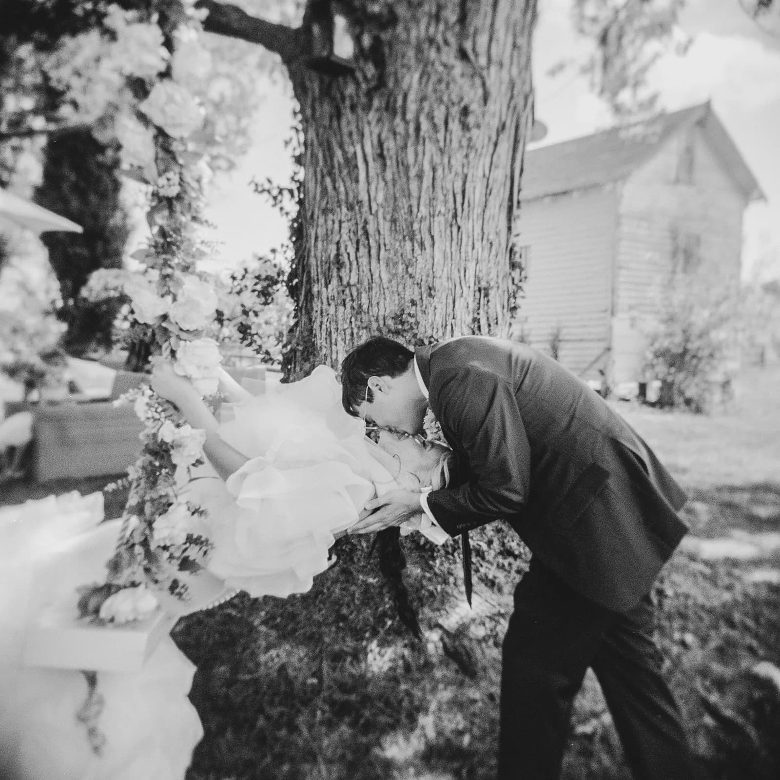 Groom dips and kisses bride beneath a large tree, surrounded by floral arch in black and white