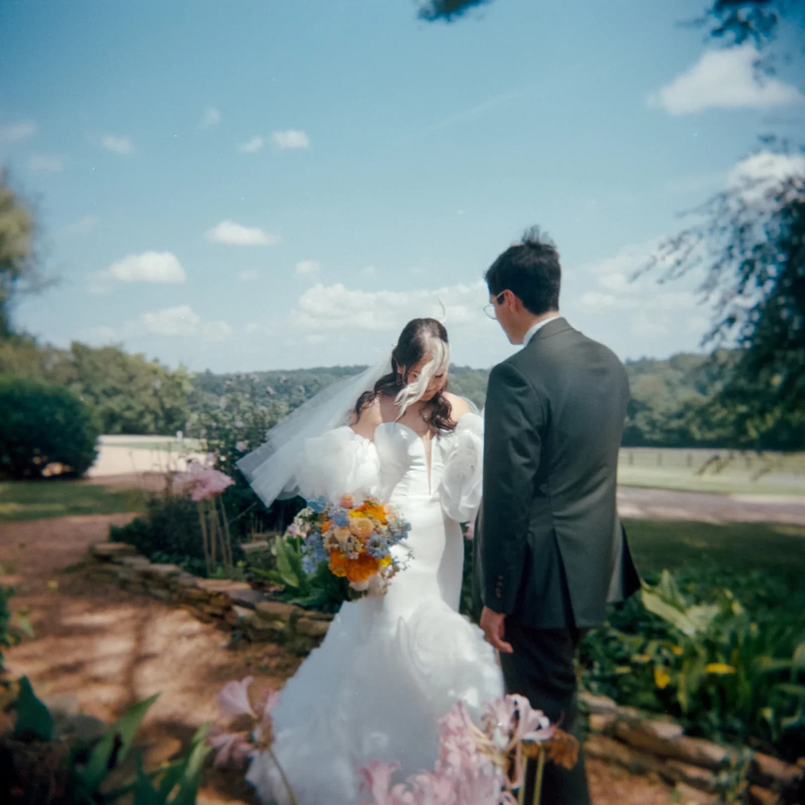 Bride and groom in wedding attire posing outdoors with yellow bouquet and scenic countryside background
