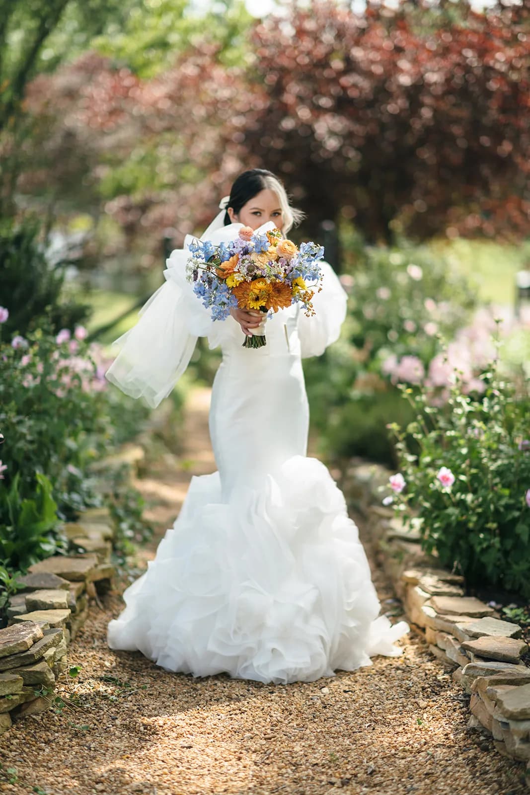 Bride in ruffled mermaid gown holding colorful bouquet along a stone garden path at Rixey Manor