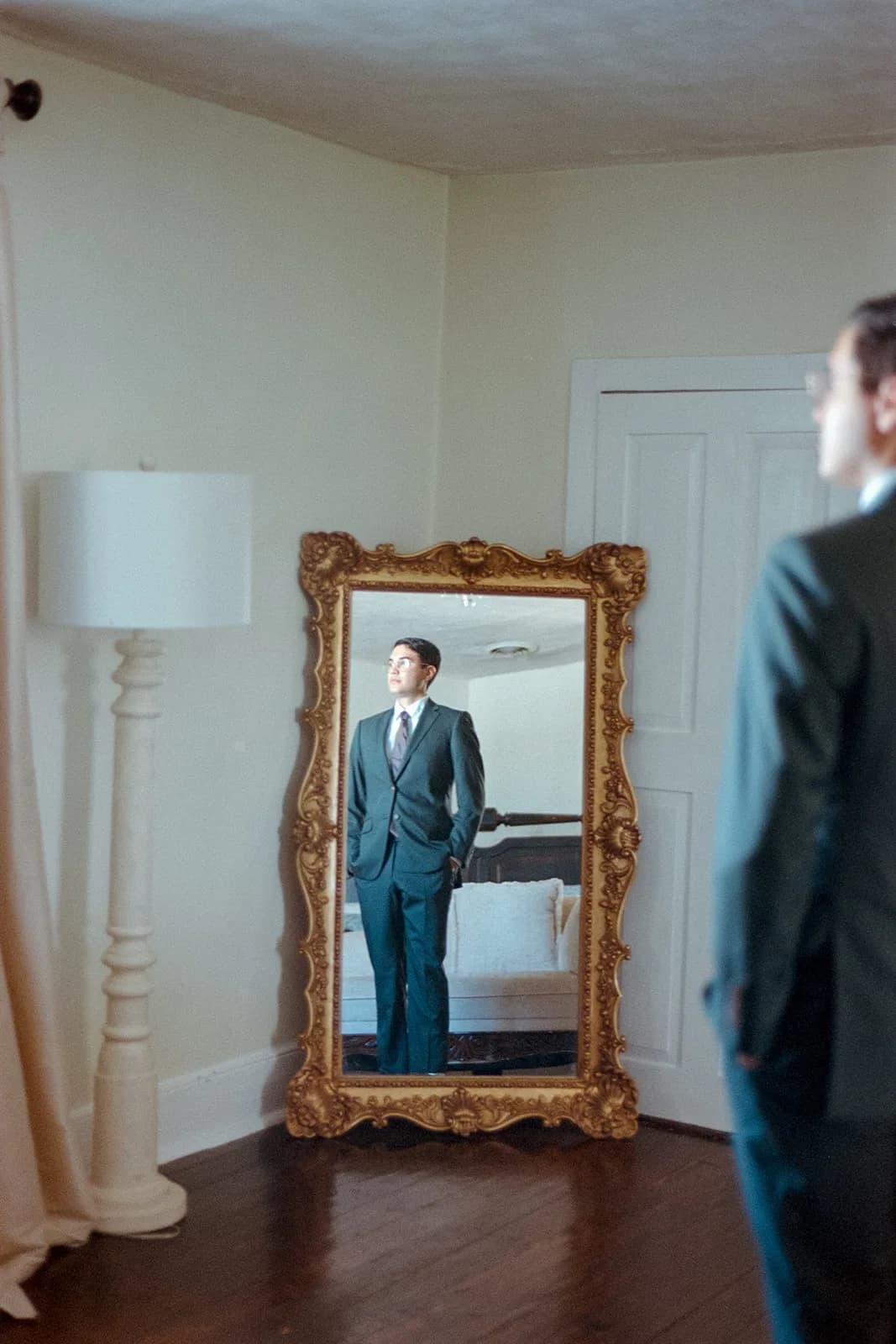 Groom in dark suit reflected in ornate gold mirror while getting ready at Rixey Manor