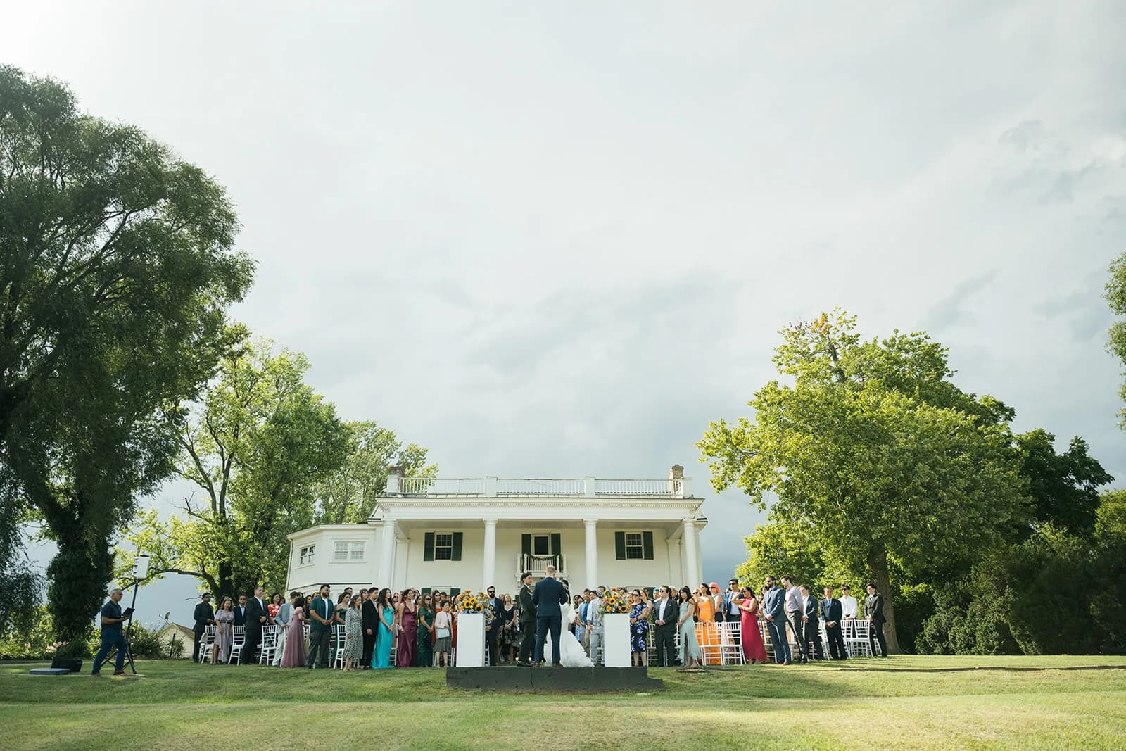 Wide-angle outdoor wedding ceremony on the lawn of a white colonial manor estate with guests gathered