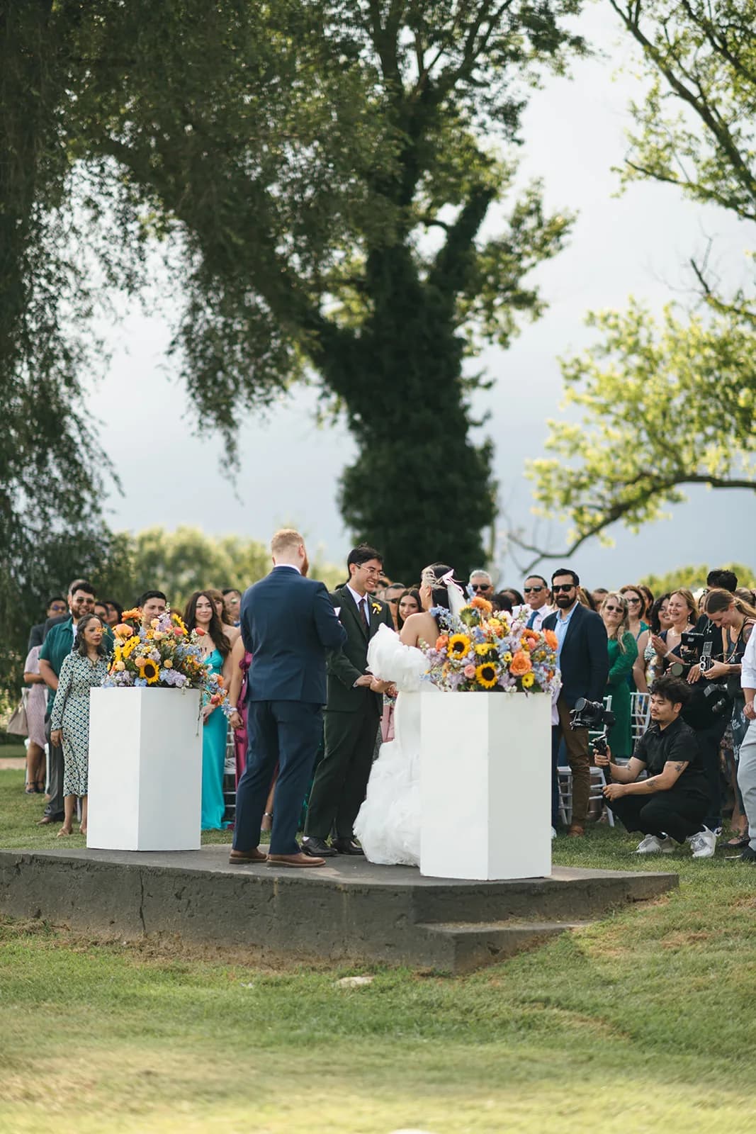 Couple exchanges vows on outdoor ceremony stage surrounded by sunflower arrangements and guests at a garden wedding