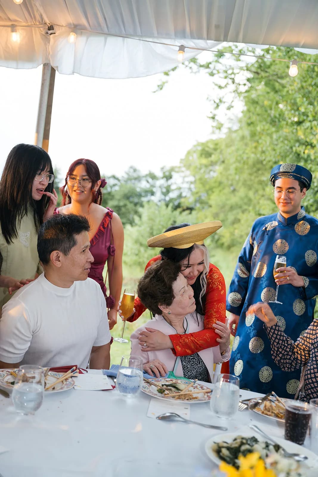 Bride in red áo dài embraces elderly guest at outdoor tented reception as family gathers around