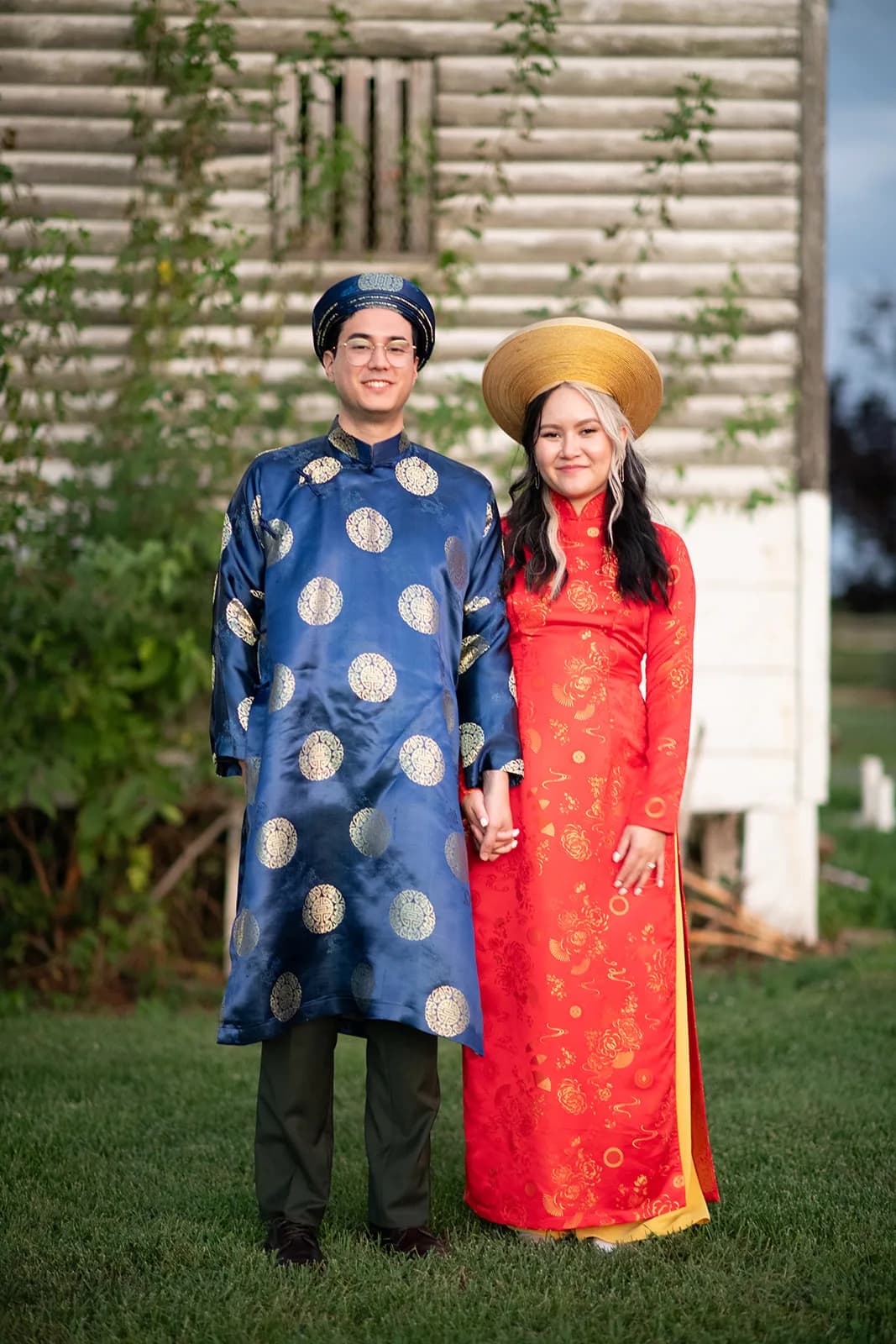 Couple in traditional Vietnamese ao dai wedding attire holding hands outdoors, smiling warmly at camera
