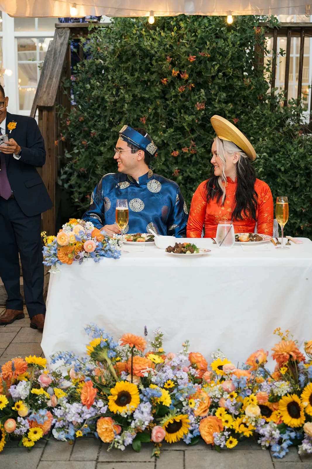 Couple in traditional Vietnamese ao dai attire laughing at sweetheart table surrounded by sunflowers and blue blooms
