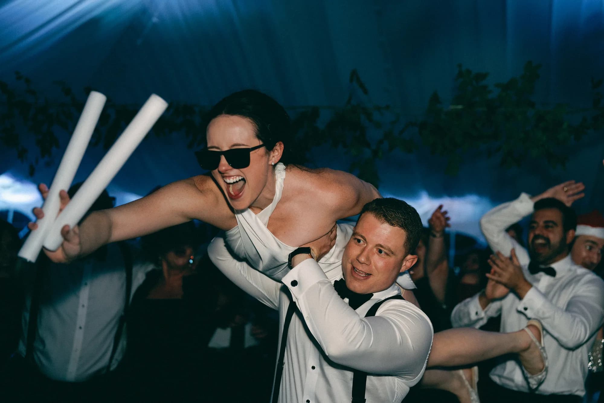 Bride carried on shoulders by groom, laughing with glow sticks on the dance floor at a lively wedding reception
