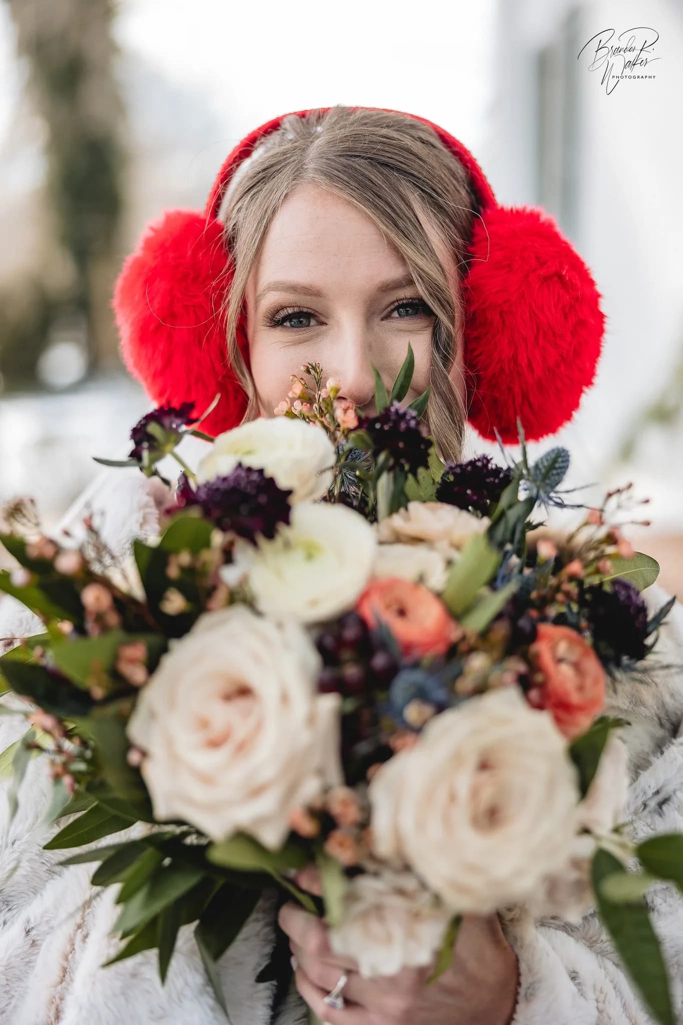 Winter bride in red earmuffs smiling over a lush bouquet of roses and dark florals