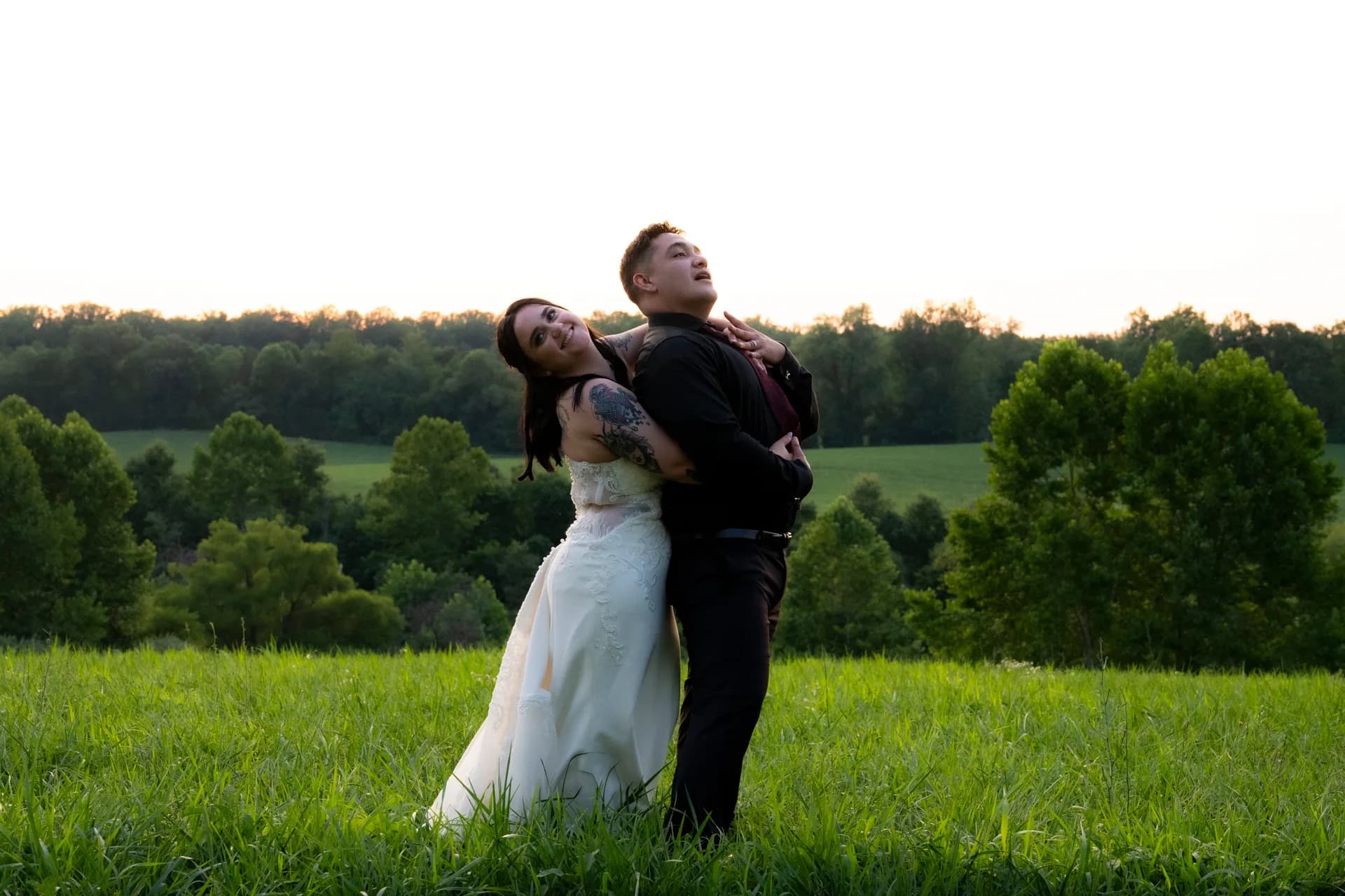 Groom lifting bride in white dress in grassy field with trees and green landscape