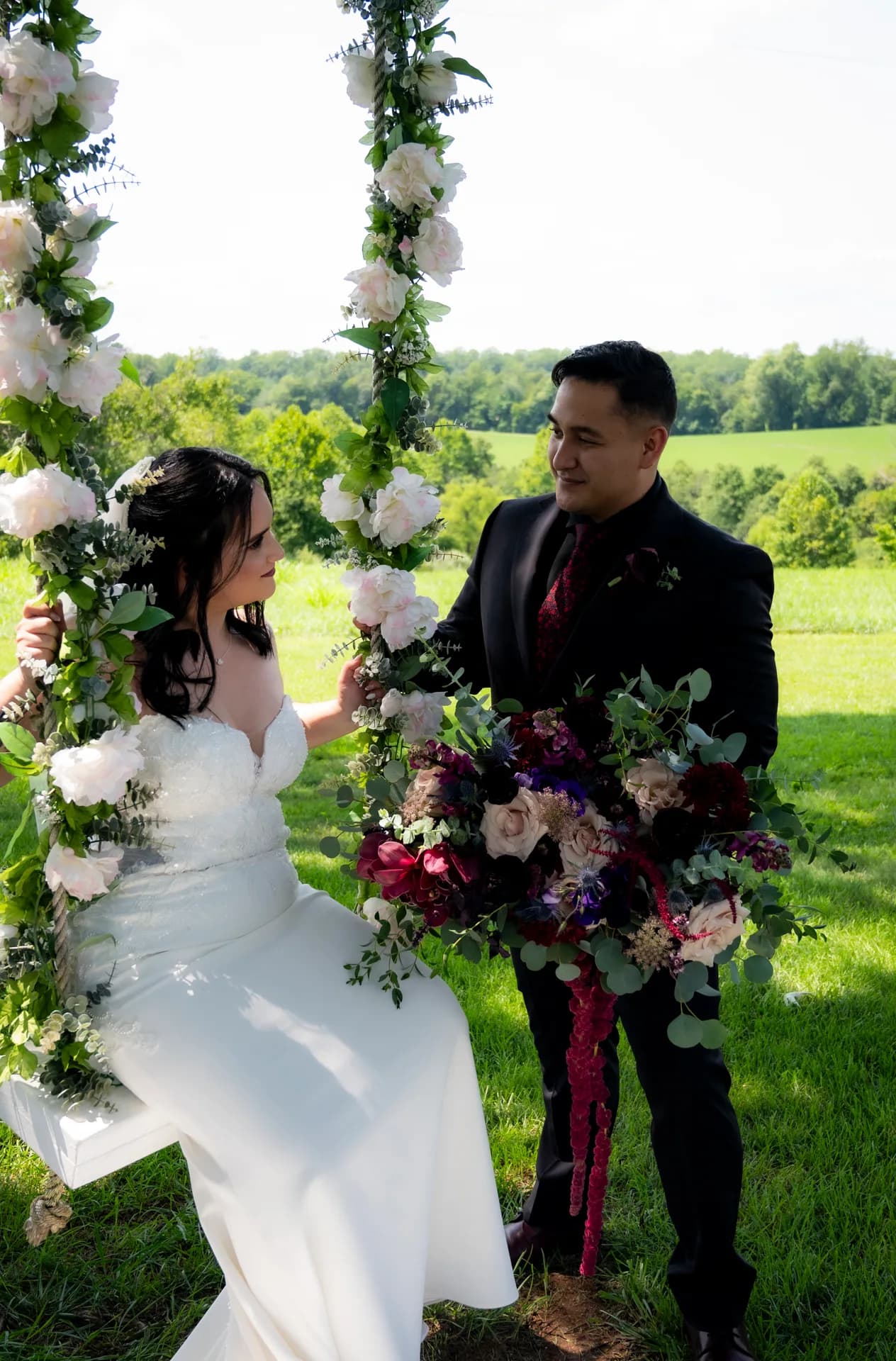 Bride on floral swing gazes at groom holding bouquet on Rixey Manor's lush green grounds