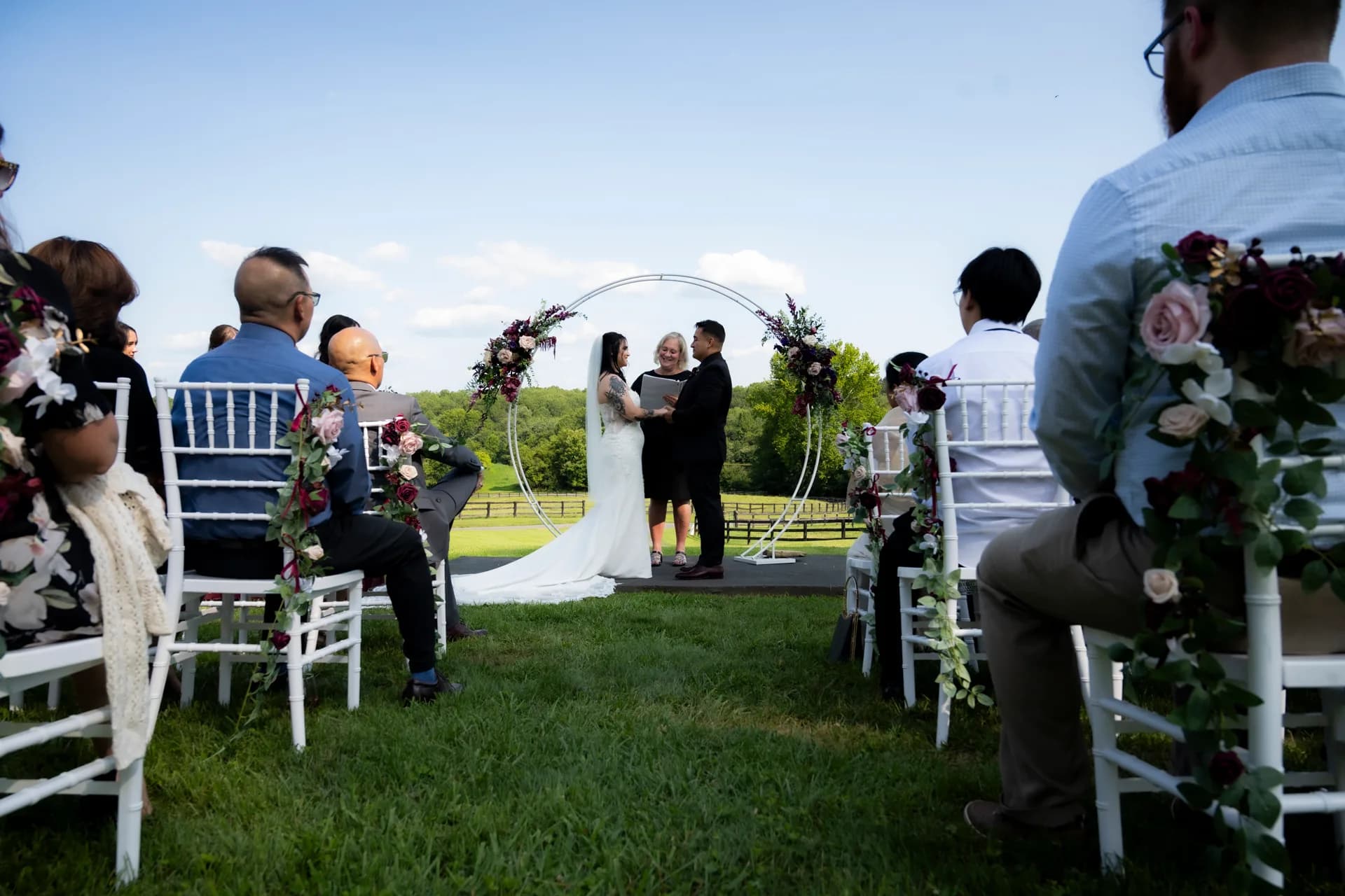 Couple exchanges vows under floral arch on Rixey Manor's green lawn as guests look on from white chairs