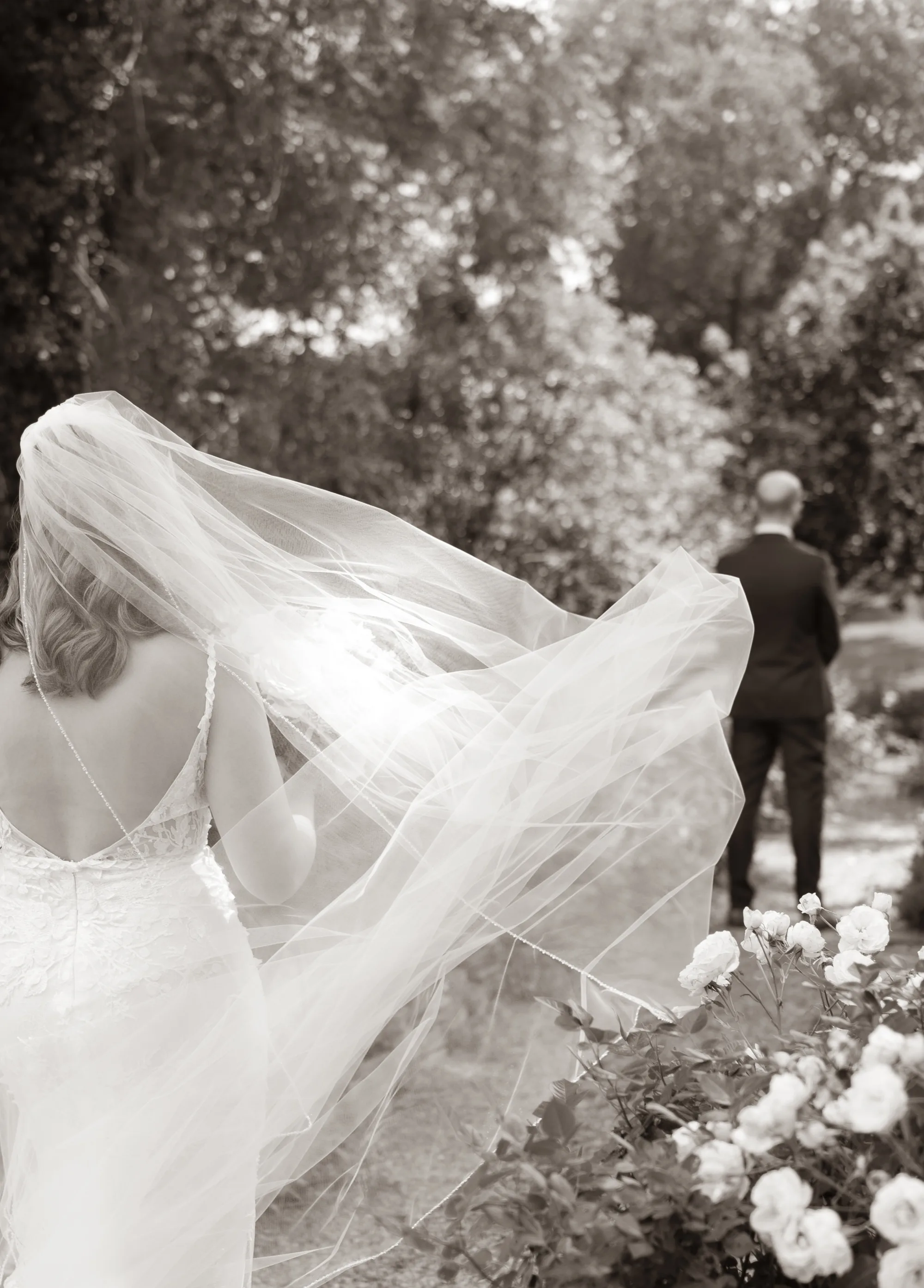 Bride's veil billows in the wind as groom walks ahead through a lush garden in black and white