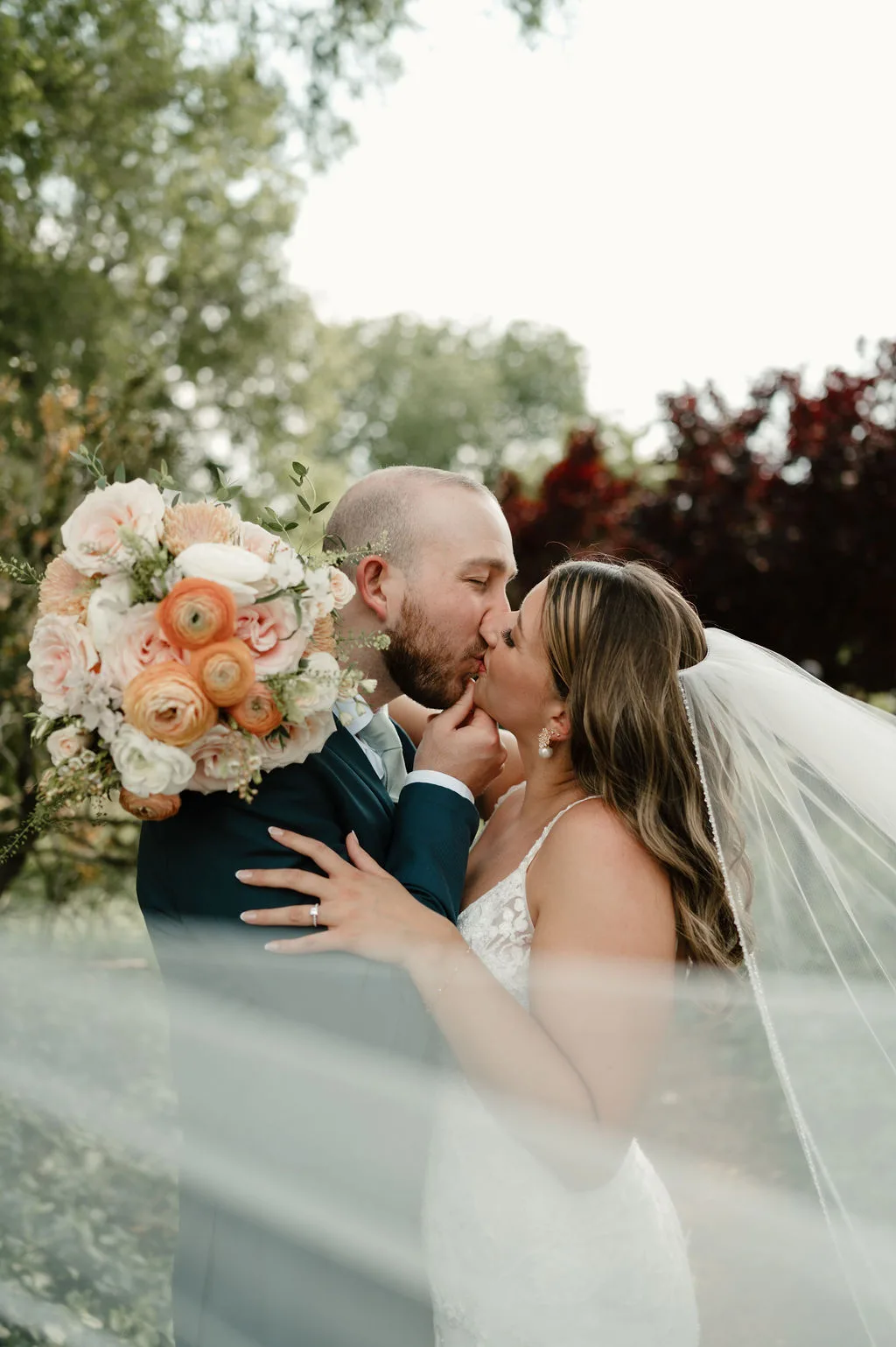 Bride and groom share a kiss beneath flowing veil, bridal bouquet of peach and blush ranunculus in foreground