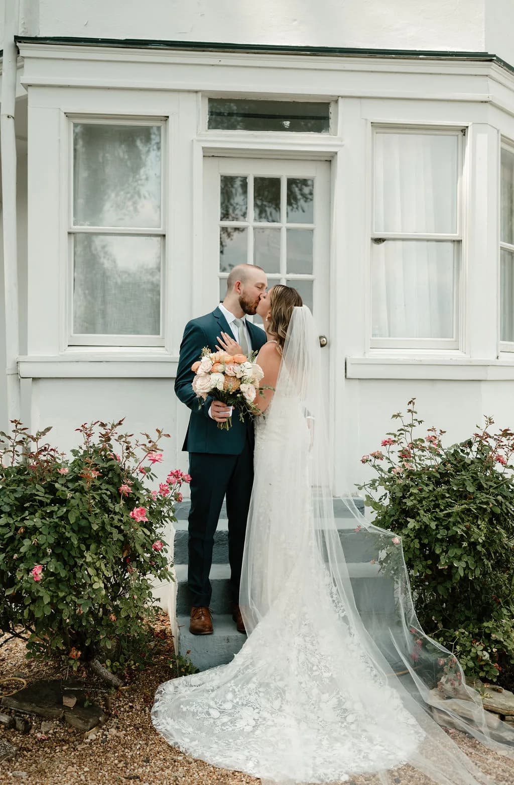 Bride and groom share a kiss on the steps of Rixey Manor's white manor house, framed by blooming rose bushes.