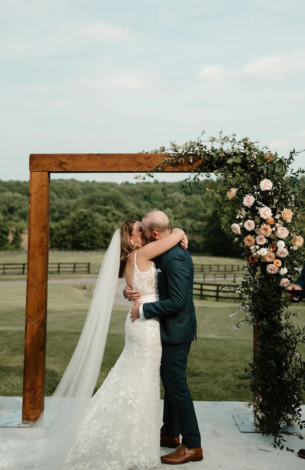 Bride and groom share first kiss under rustic wood arch with floral greenery at Rixey Manor outdoor ceremony