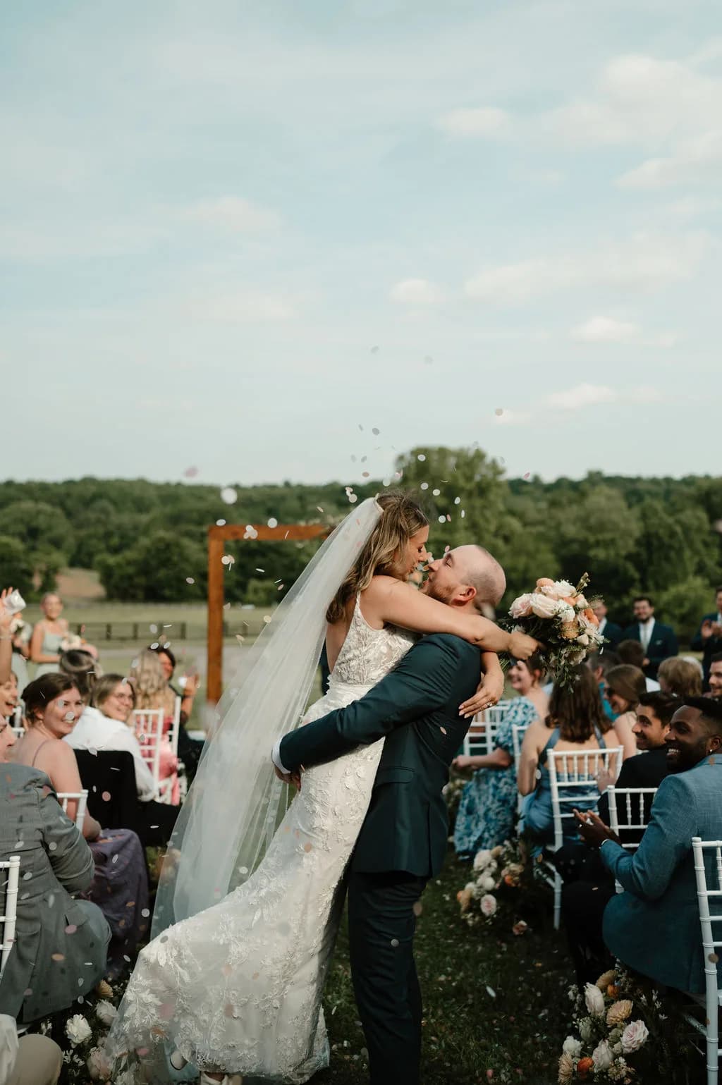 Groom lifts bride for a kiss at outdoor ceremony aisle as guests toss confetti on lush green grounds