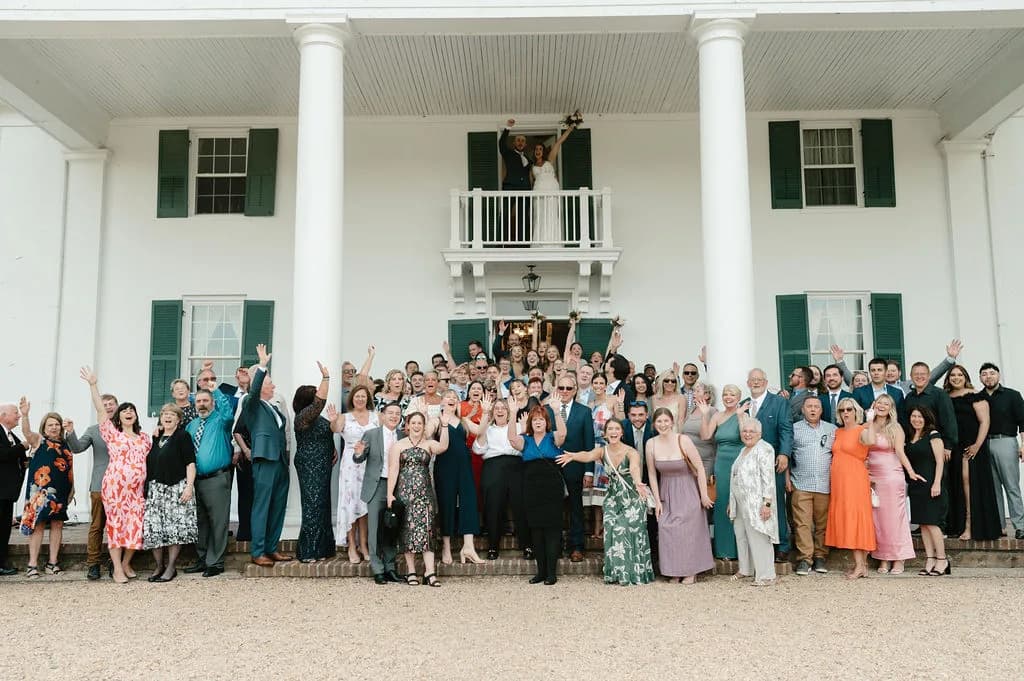 Joyful wedding group photo on Rixey Manor's columned front steps, couple waving from balcony above