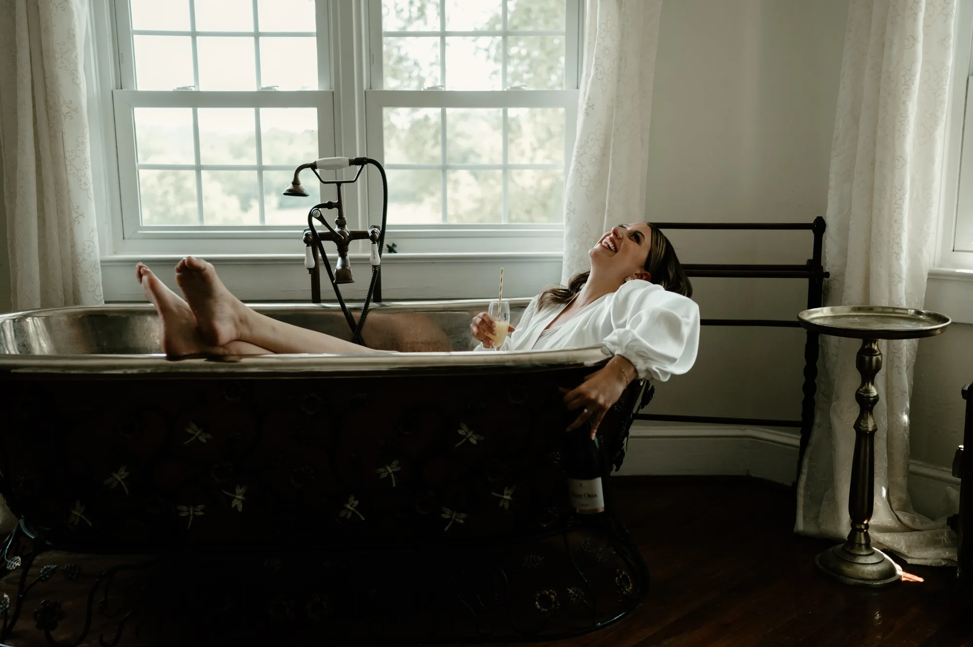 Bride laughing in a clawfoot bathtub in a white robe, holding a glass, in a vintage bathroom