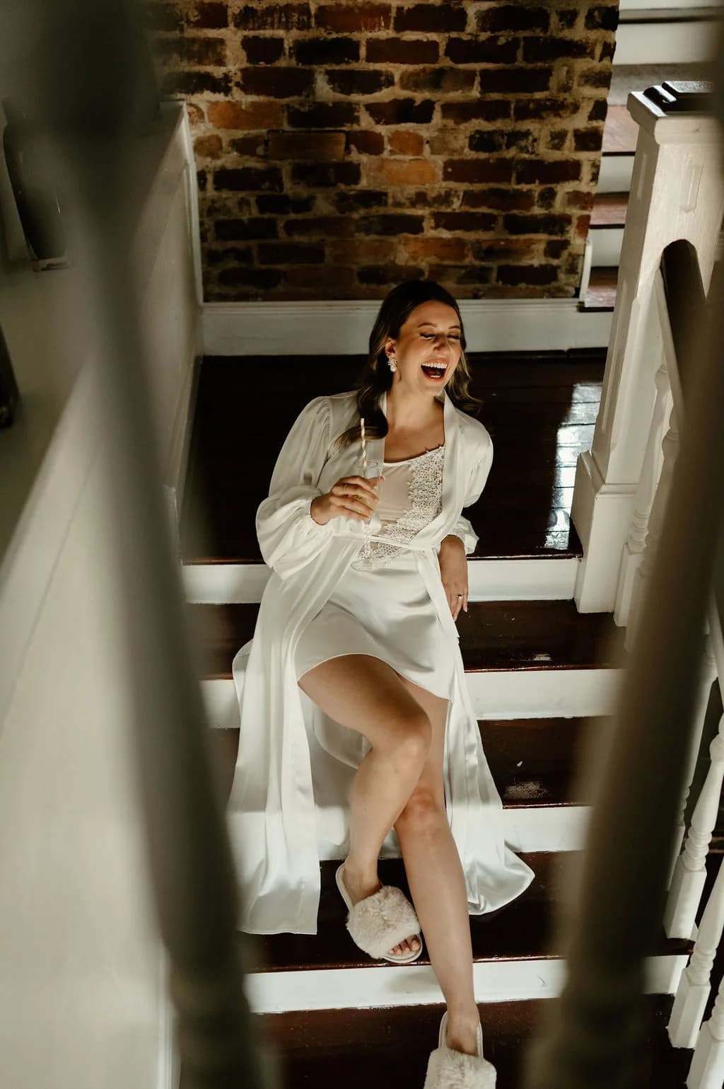 Bride in white robe laughing on staircase with exposed brick wall behind her