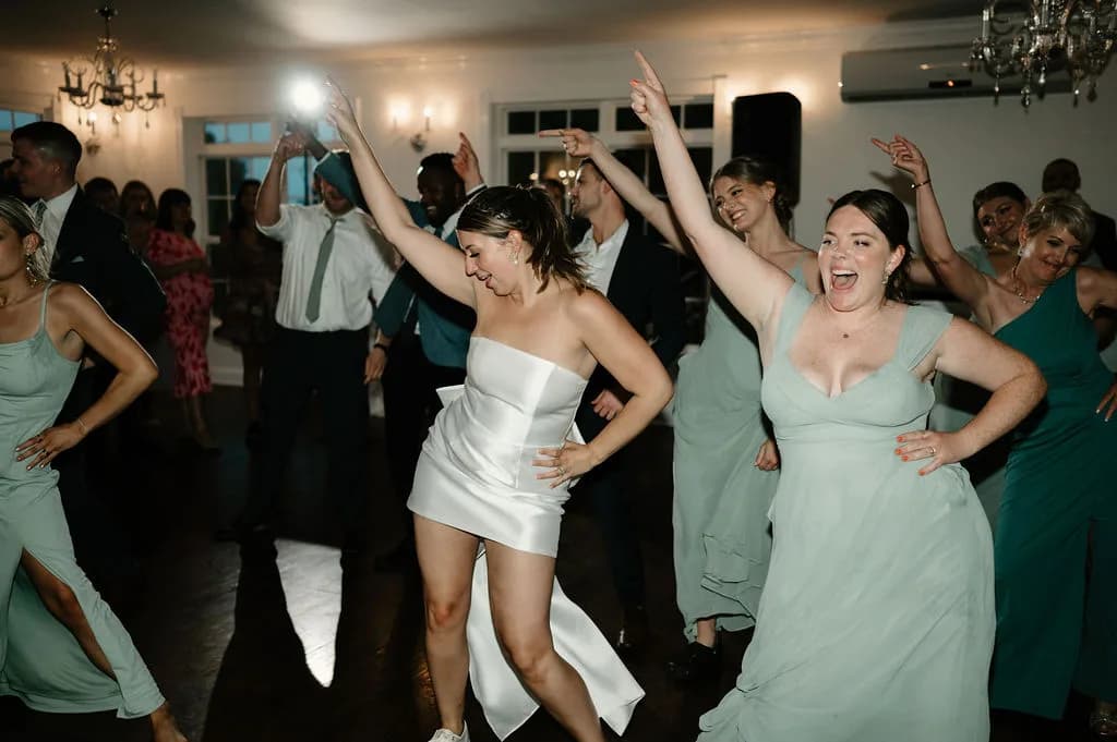 Bride in short white dress dancing joyfully with bridesmaids in sage green at wedding reception