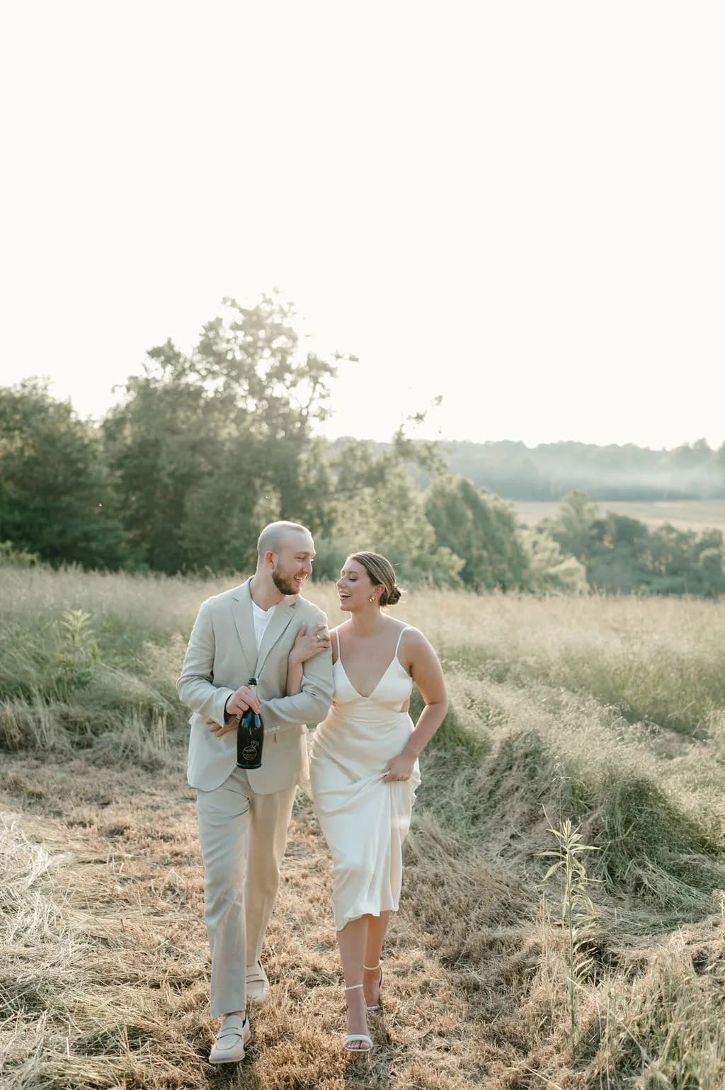 Couple laughing together while walking through a golden sunlit meadow, groom holding a champagne bottle