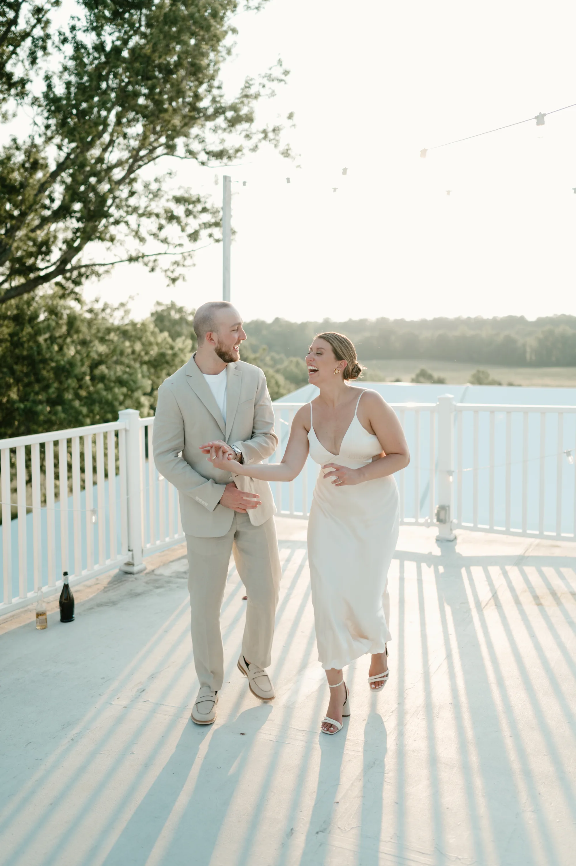 Couple in light neutral wedding attire walking on a sunny deck overlooking water