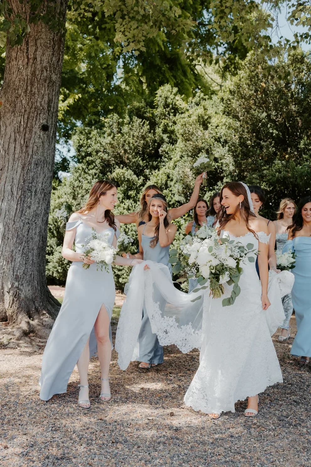Bride laughing with bridesmaids in dusty blue gowns walking outdoors under large trees