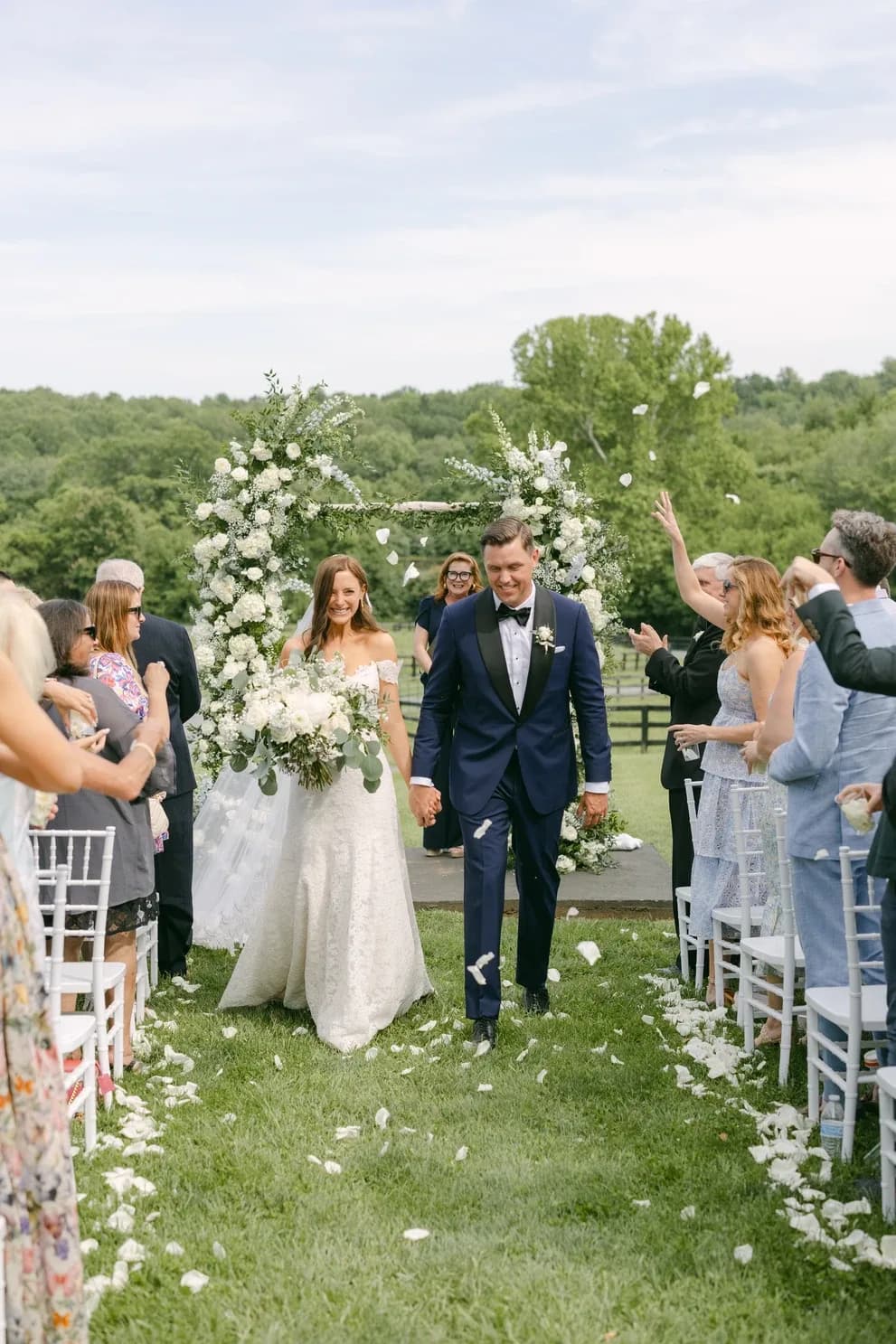 Newlyweds walk the aisle at Rixey Manor outdoor ceremony as guests toss petals beneath a white floral arch