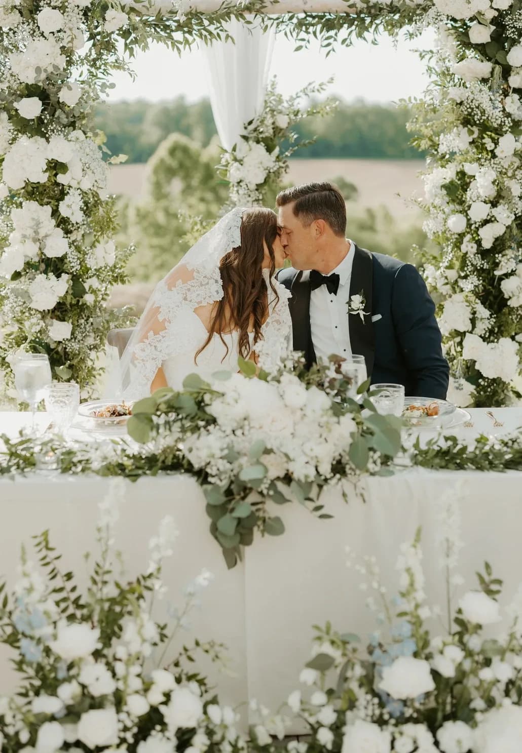 Bride and groom share a kiss at sweetheart table framed by white floral arch with Virginia countryside backdrop