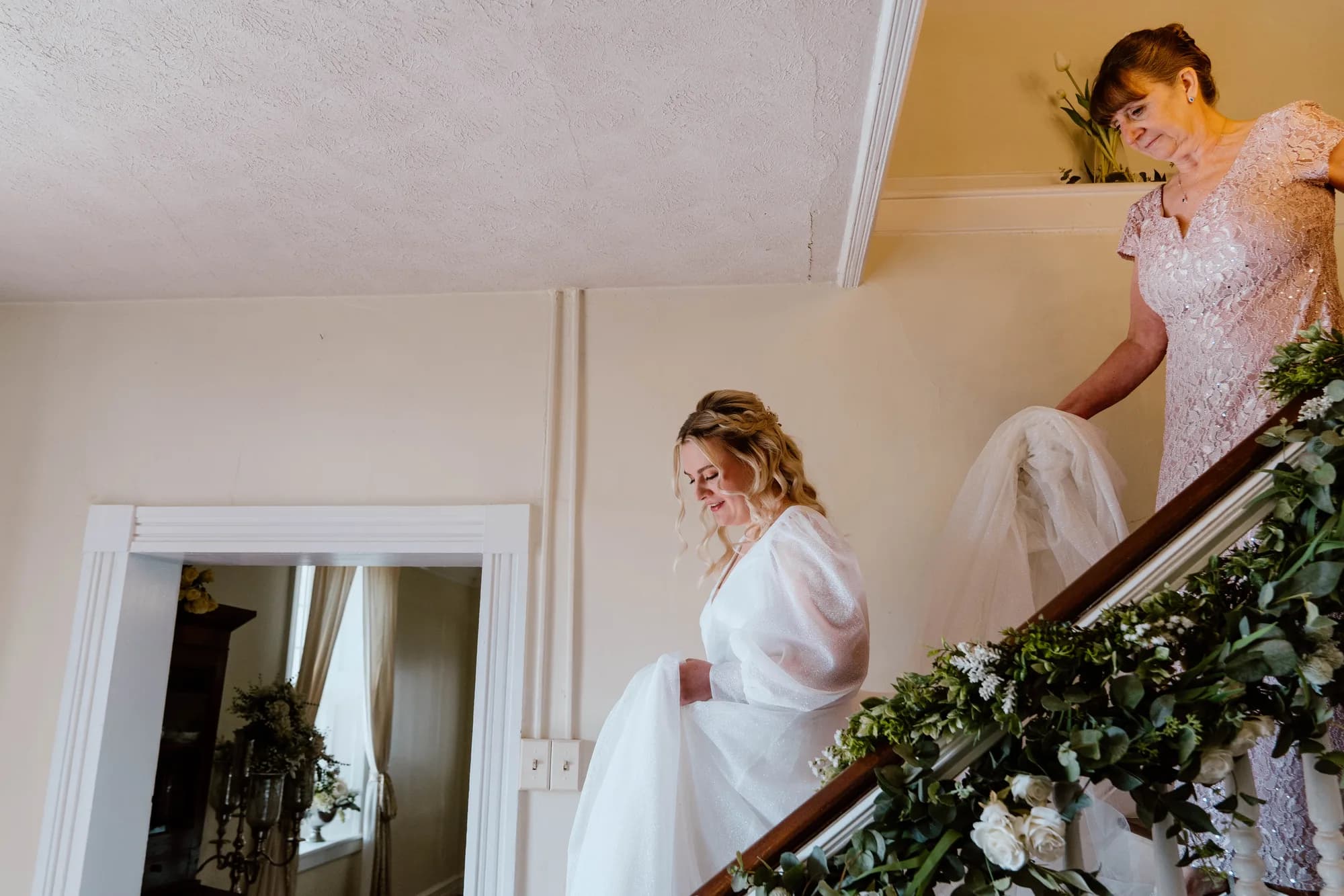Bride descends floral-adorned staircase at Rixey Manor as attendant helps carry her gown train