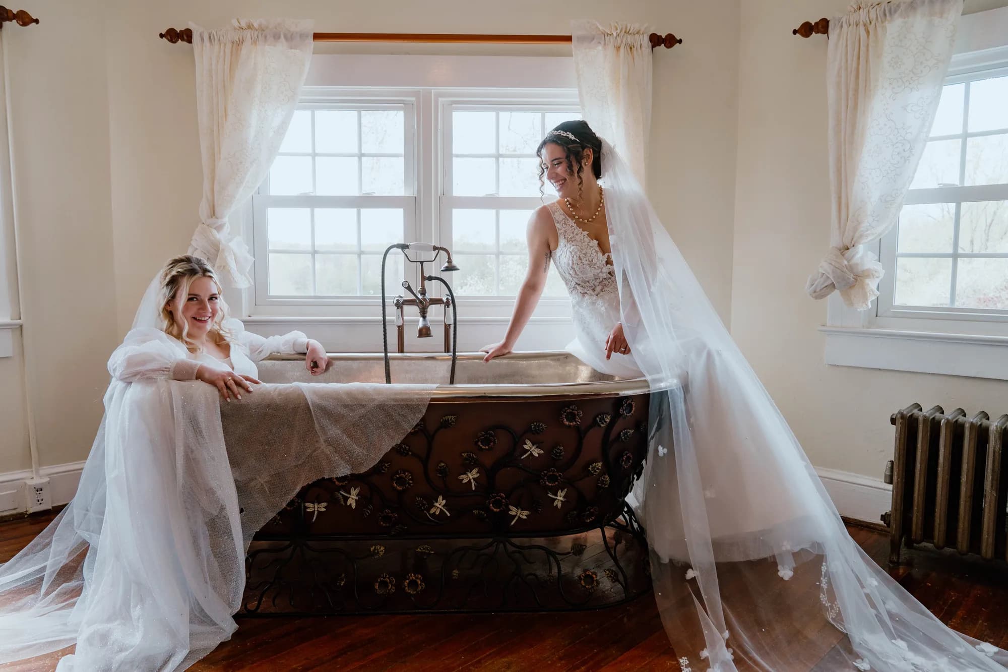 Two brides in wedding gowns laughing beside an ornate vintage clawfoot tub in a sunlit Rixey Manor suite