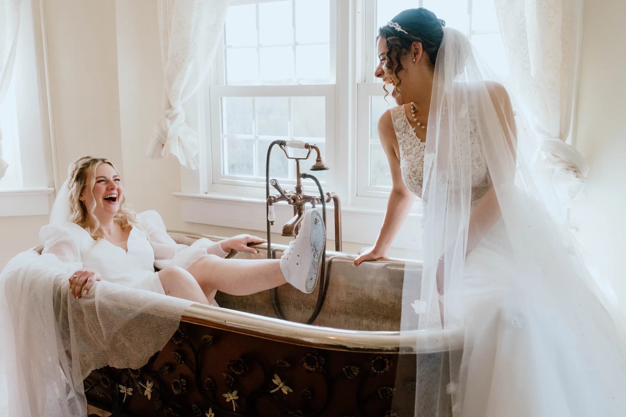 Two brides laughing together in a vintage clawfoot tub during getting-ready moments at Rixey Manor