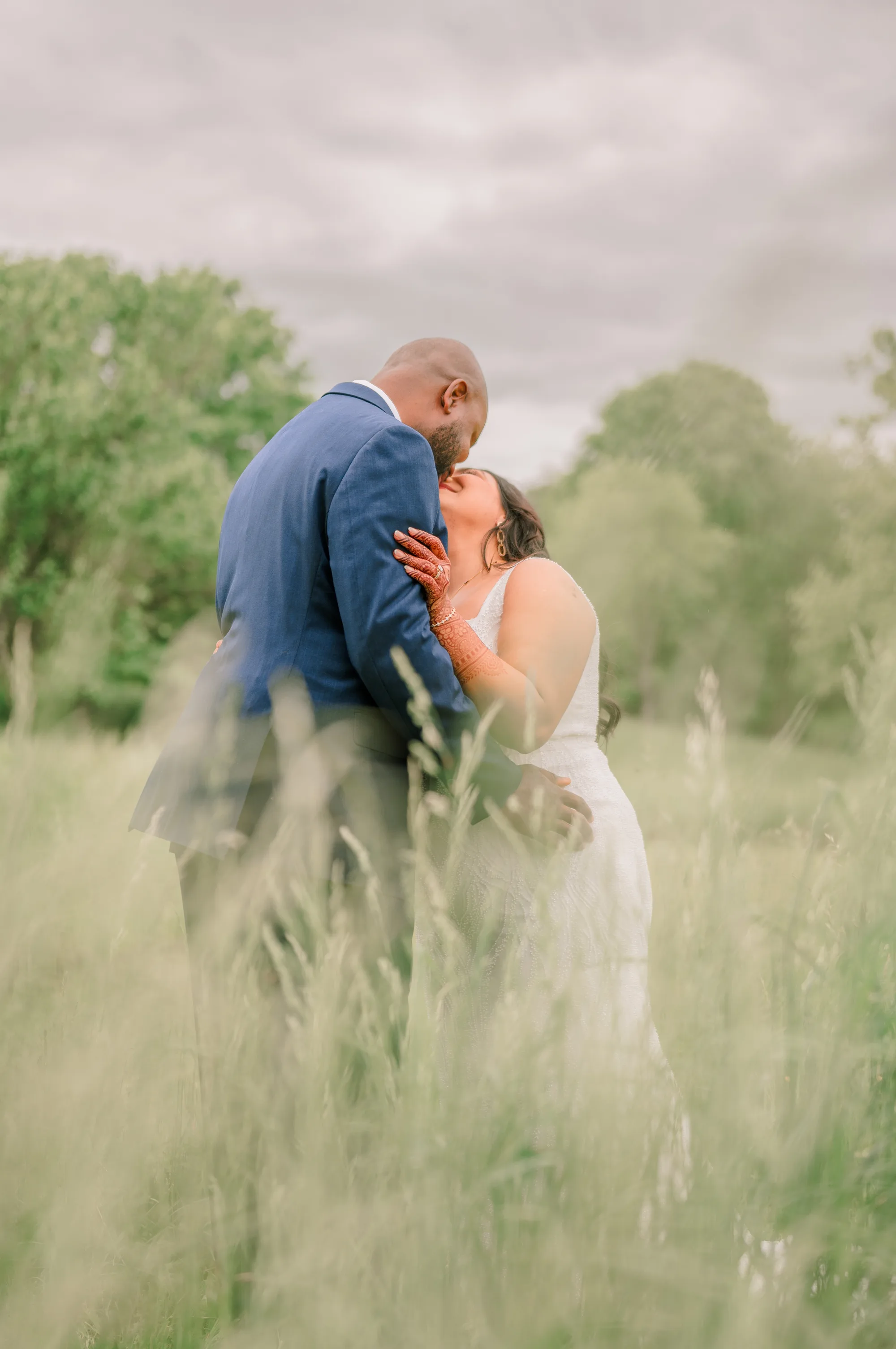 Couple kissing in tall grass field with trees and cloudy sky in background