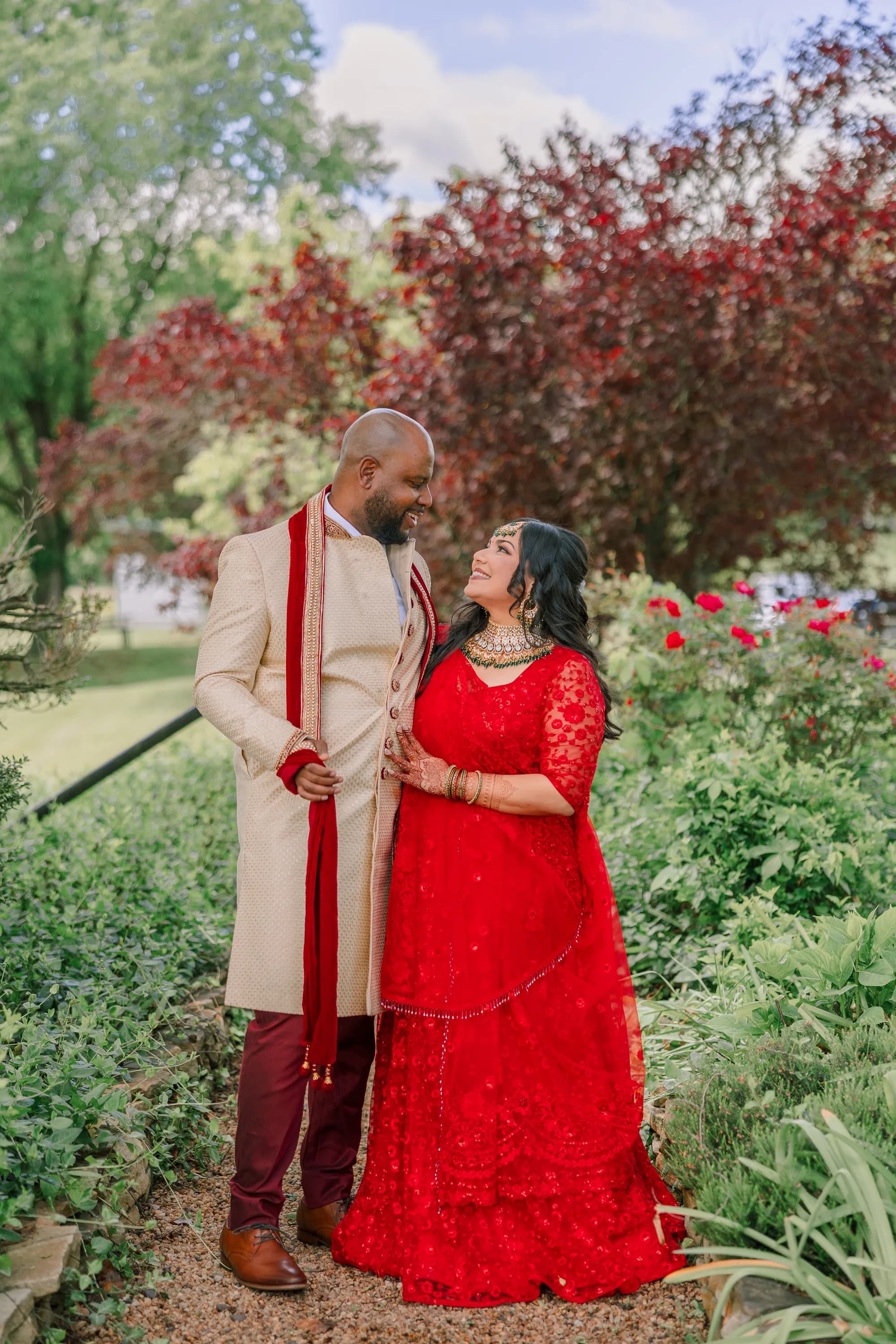Bride in red lehenga and groom in cream sherwani share a joyful gaze in Rixey Manor's lush garden