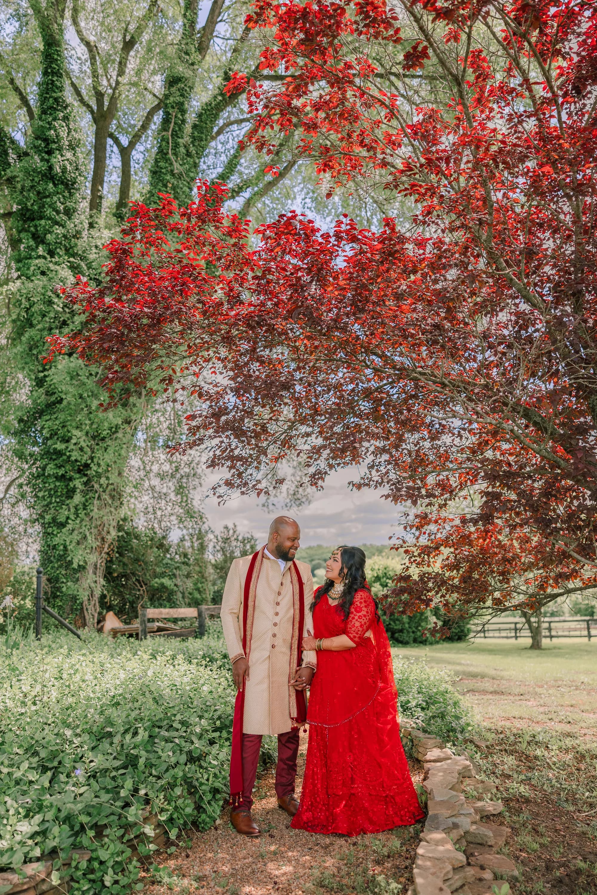 Couple in red and gold Indian wedding attire holding hands under a crimson tree on Rixey Manor grounds