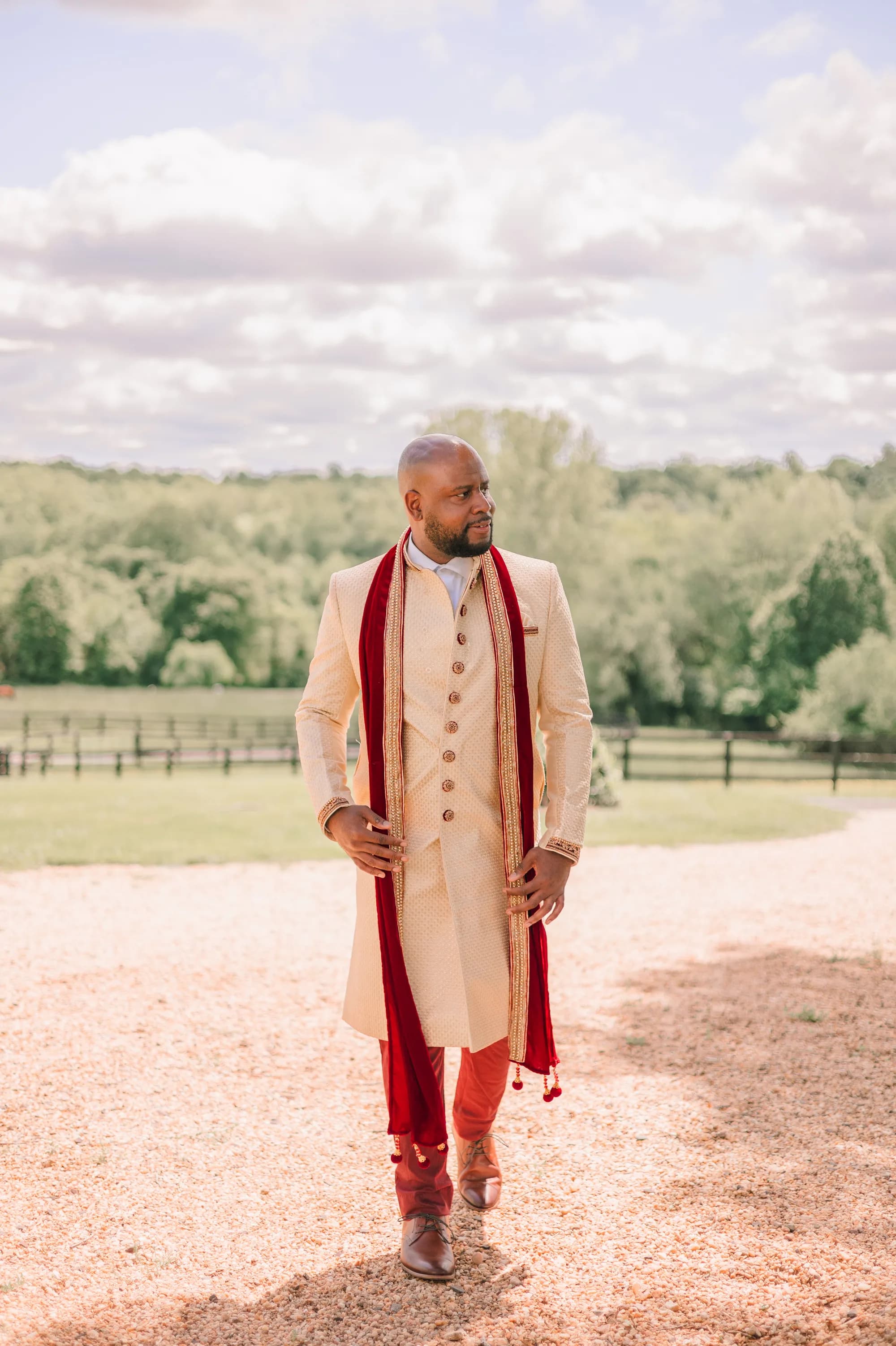 Groom in ivory sherwani with red dupatta walks Rixey Manor's gravel drive, lush grounds behind him