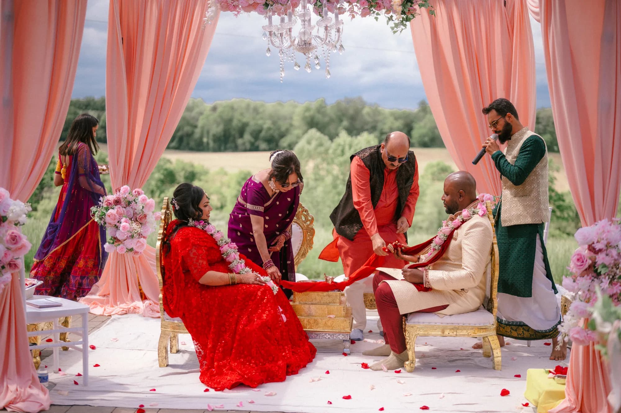Couple in vibrant traditional attire during an outdoor South Asian wedding ceremony under a pink floral mandap