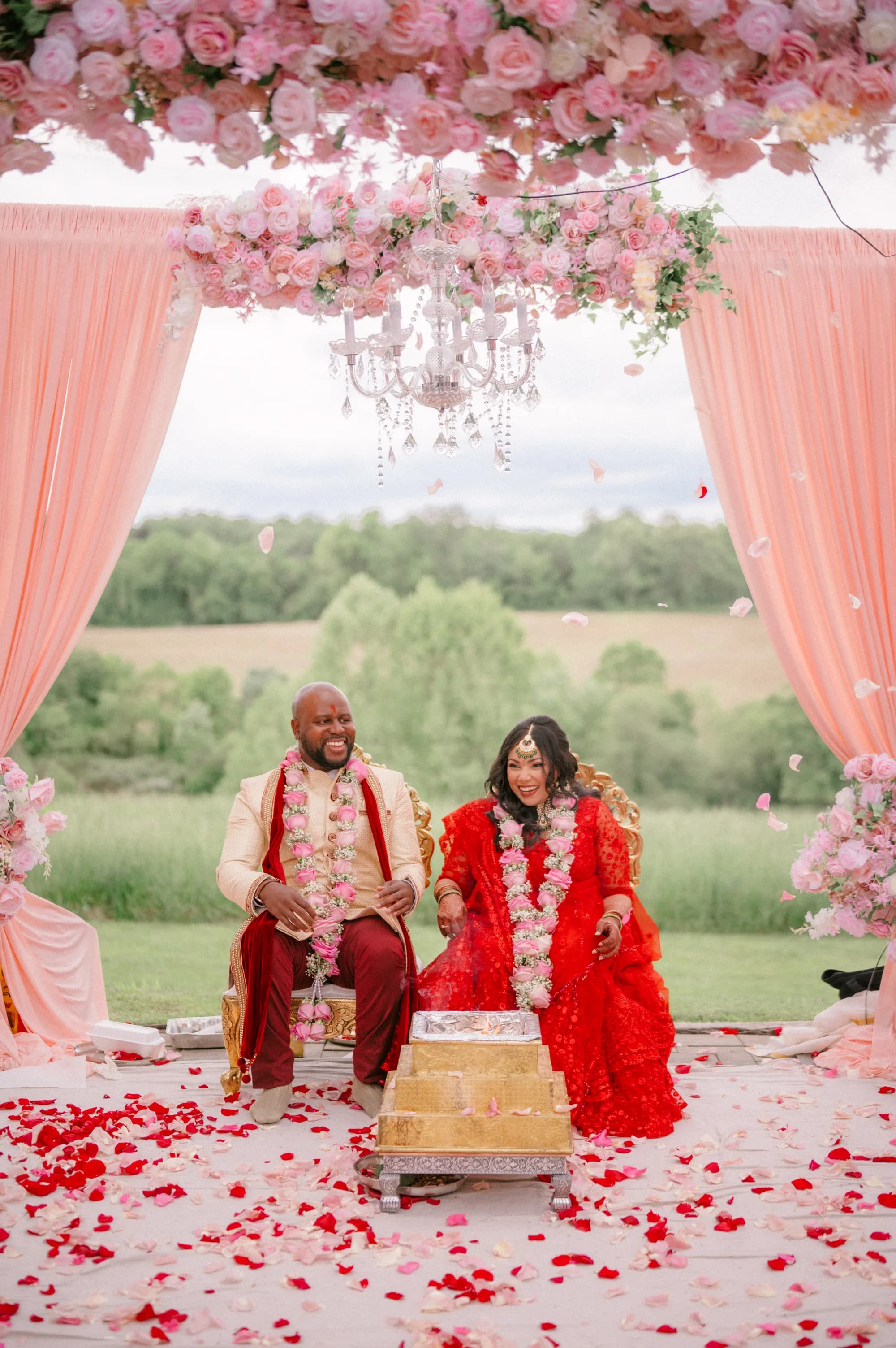 Bride and groom in traditional Indian wedding attire seated under ornate pink floral archway at ceremony