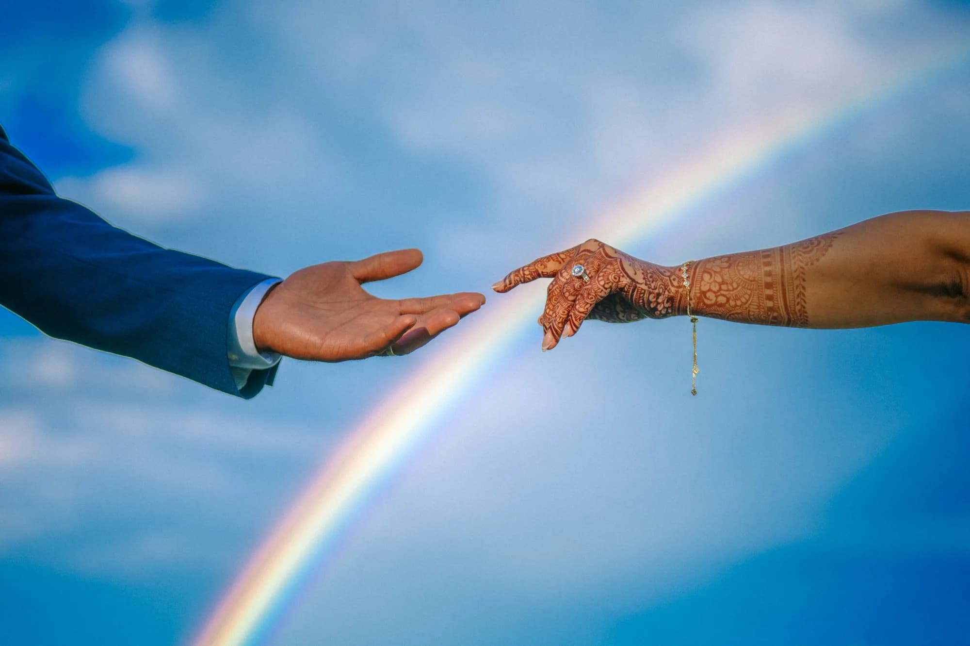 Bride's henna-adorned hand reaches toward groom's hand against a vivid blue sky with rainbow