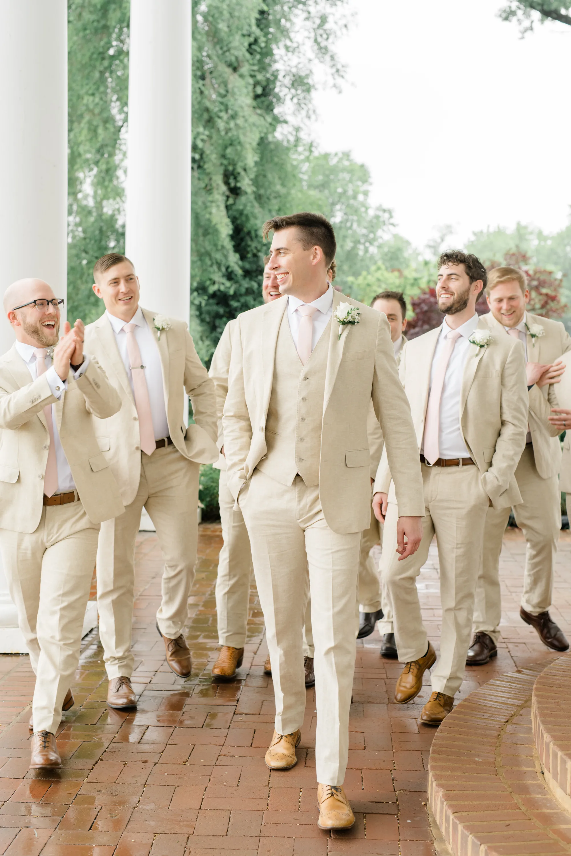 Groom and groomsmen walking and laughing under white columns at Rixey Manor in matching cream suits